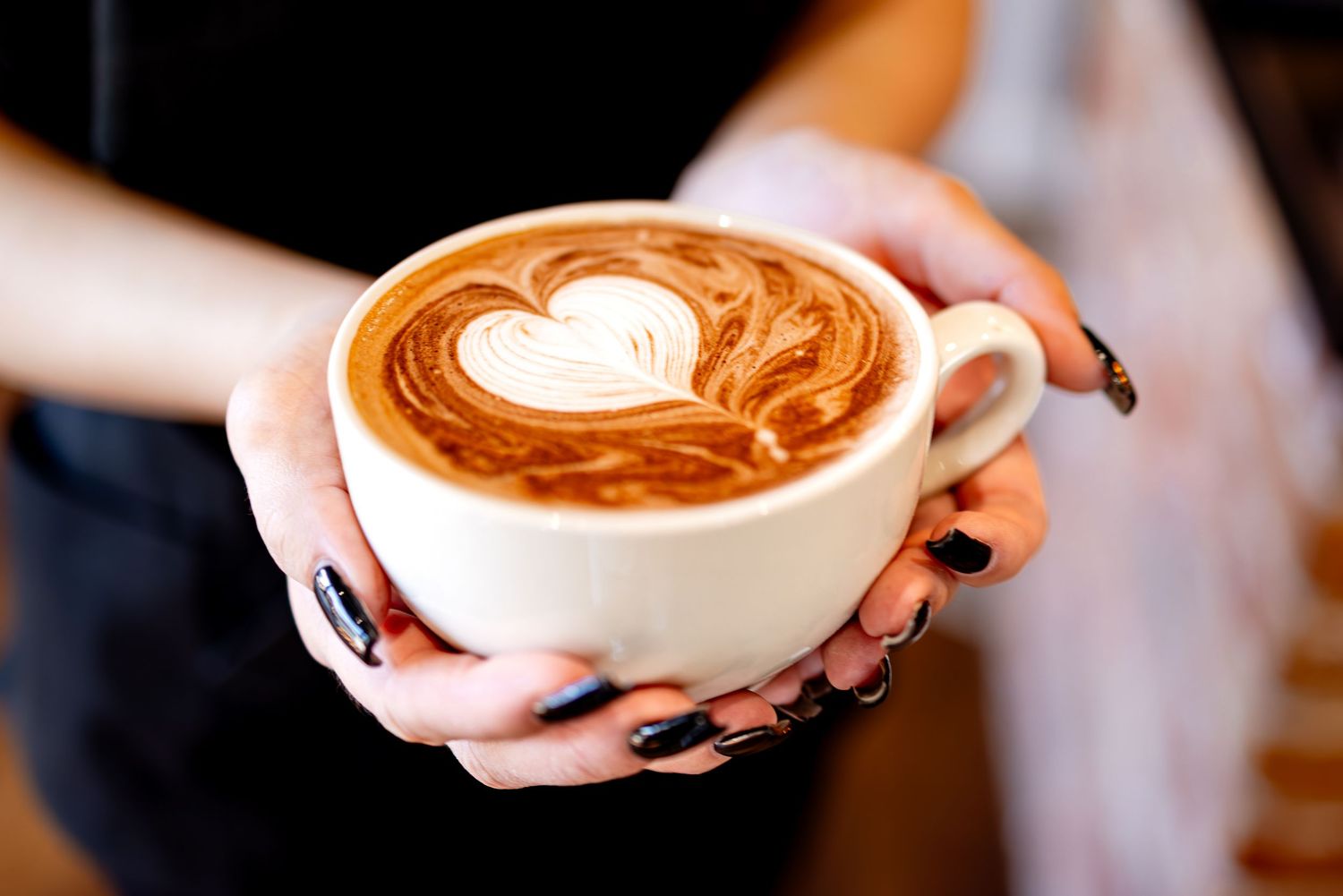 Barista holding white ceramic cup of latte art with heart design and dark nail polish.