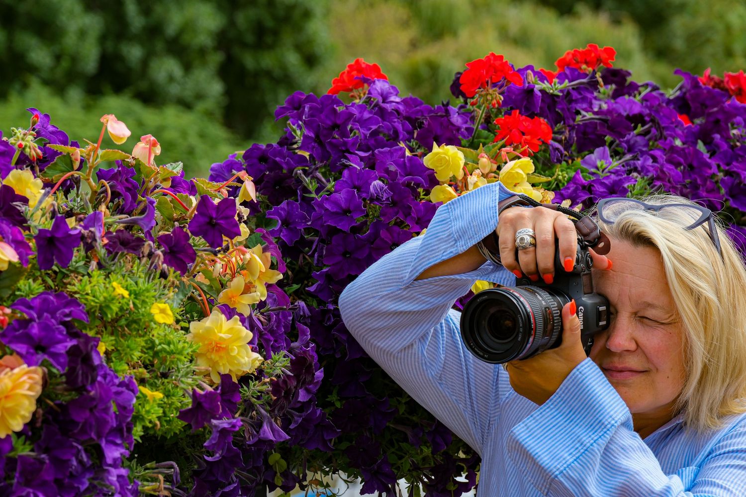 Blonde woman photographing vibrant purple, red, and yellow flowers with a Canon DSLR camera outdoors.