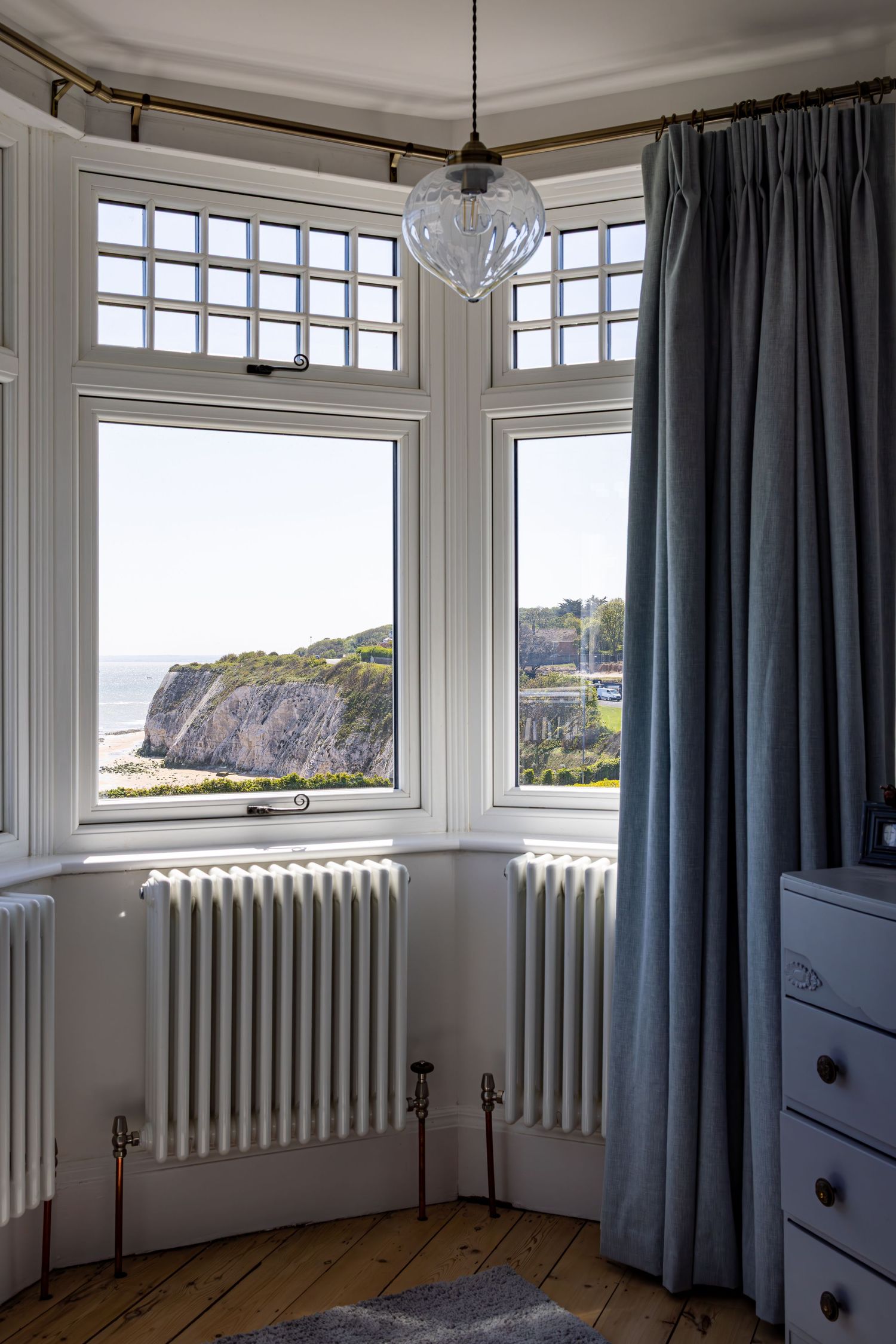 Elegant bay window bedroom with sea cliff views, white radiator, blue curtains and pendant light.