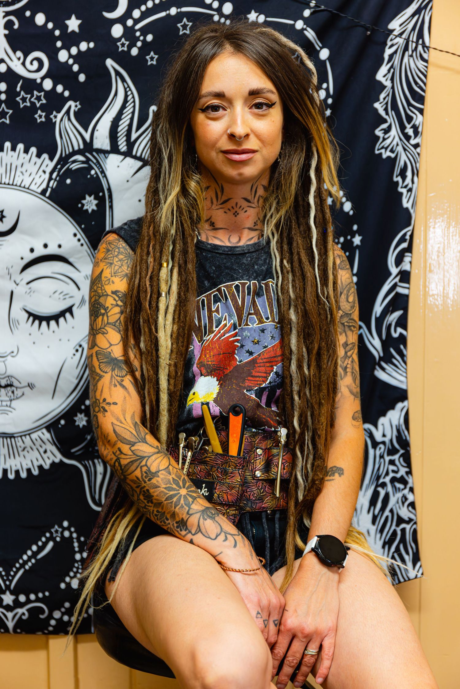 Tattooed woman with dreadlocks sitting before celestial tapestry wearing graphic tee.