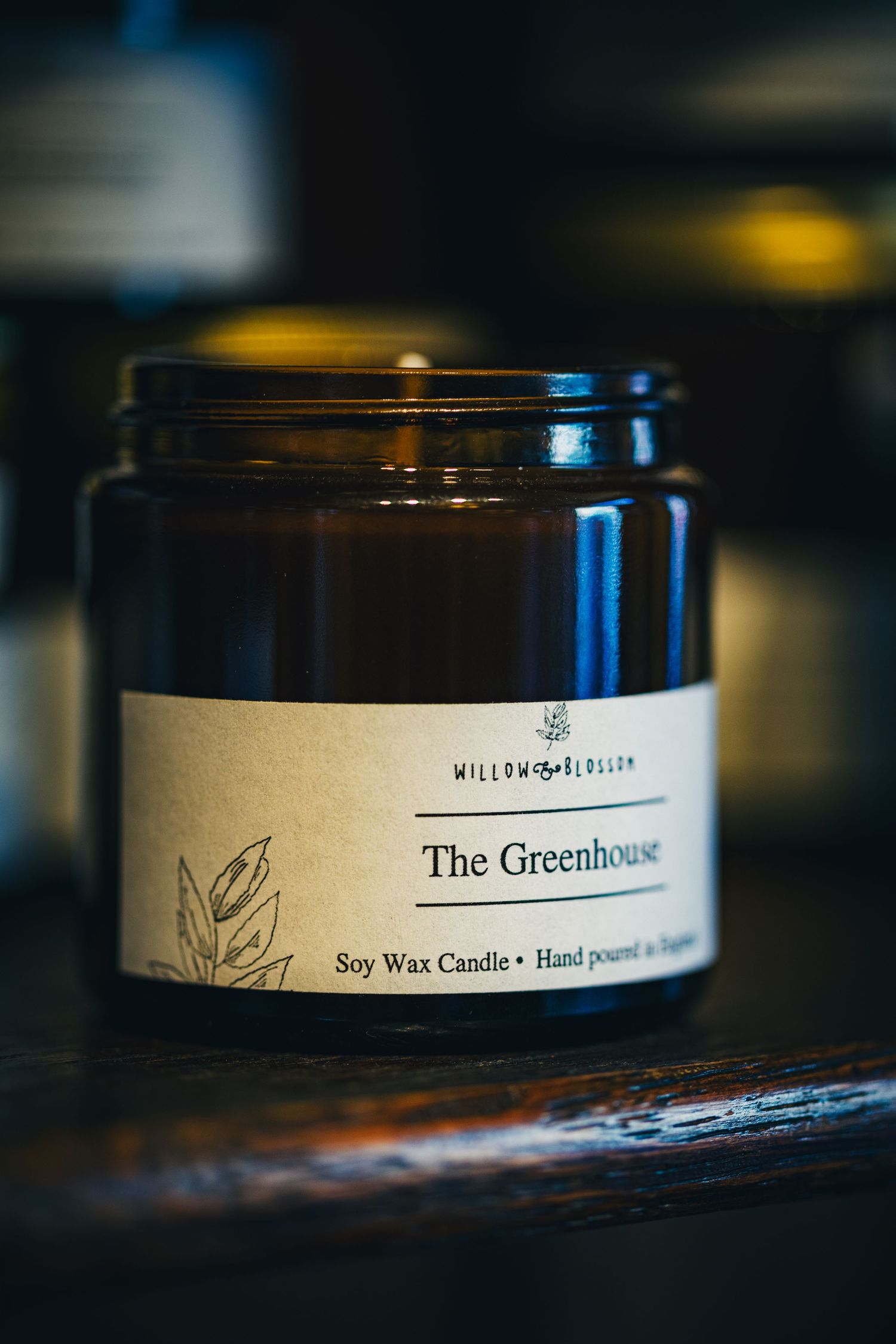 Willow Blossom soy wax candle in amber glass jar labeled The Greenhouse on wooden surface.