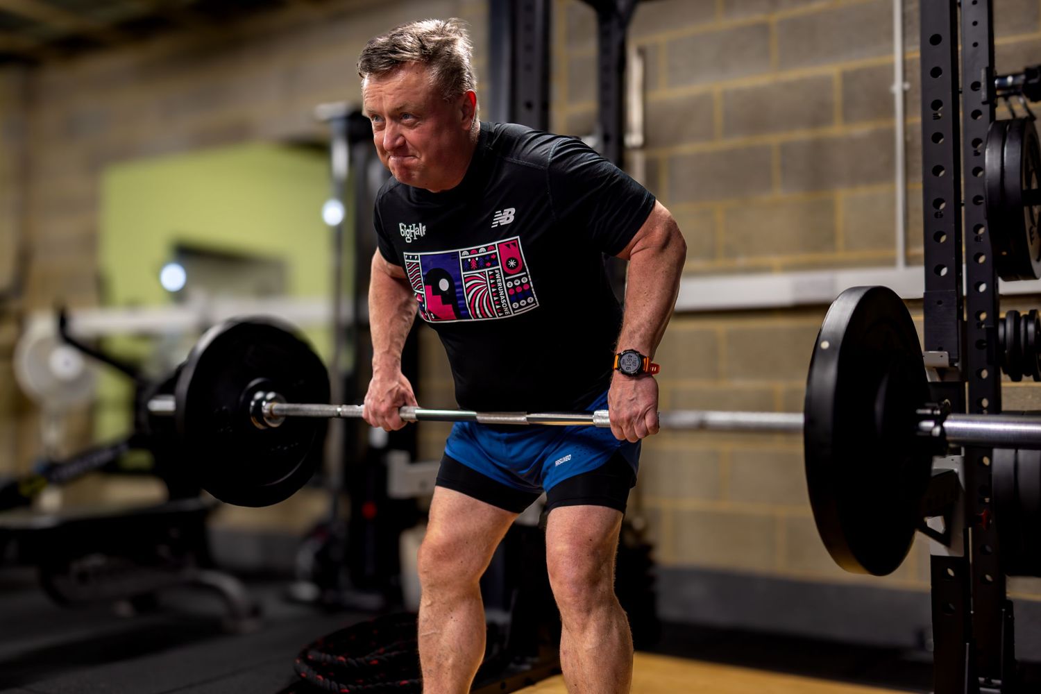 Muscular man performing a deadlift with heavy barbell in a gym, wearing New Balance shirt and blue shorts.