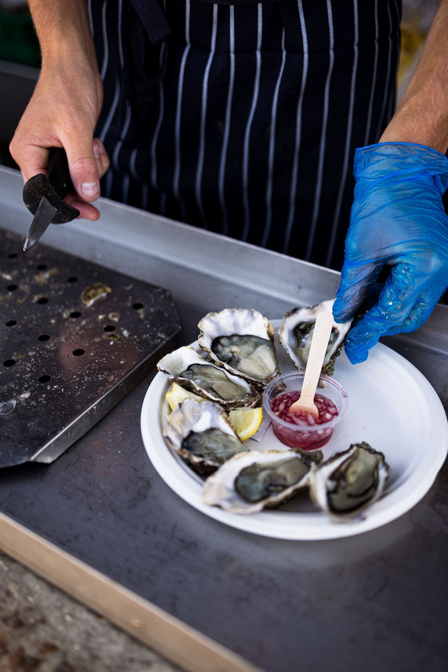 Chef in blue gloves shucking fresh oysters on a white plate with lemon and sauce.