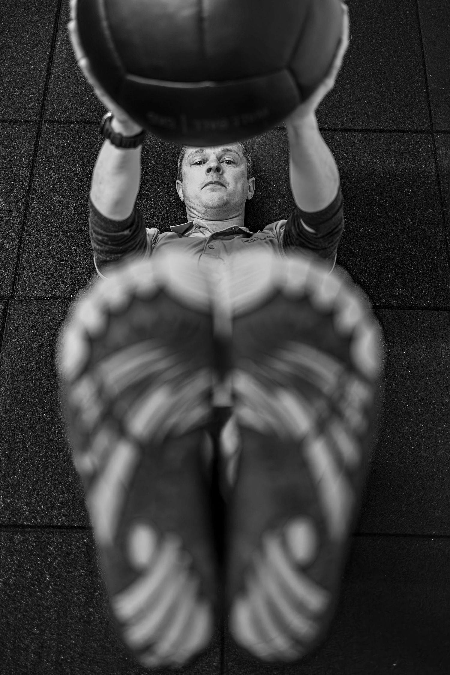 Black and white photo of a man lying on gym floor doing medicine ball chest press exercise viewed from above.