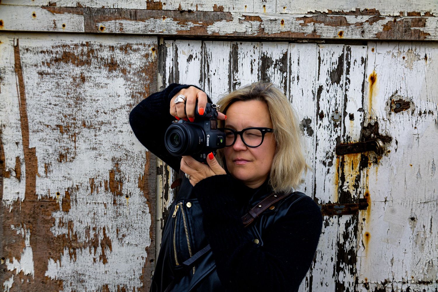 Female photographer with blonde hair and red glasses holding a camera outdoors near a rustic wall.