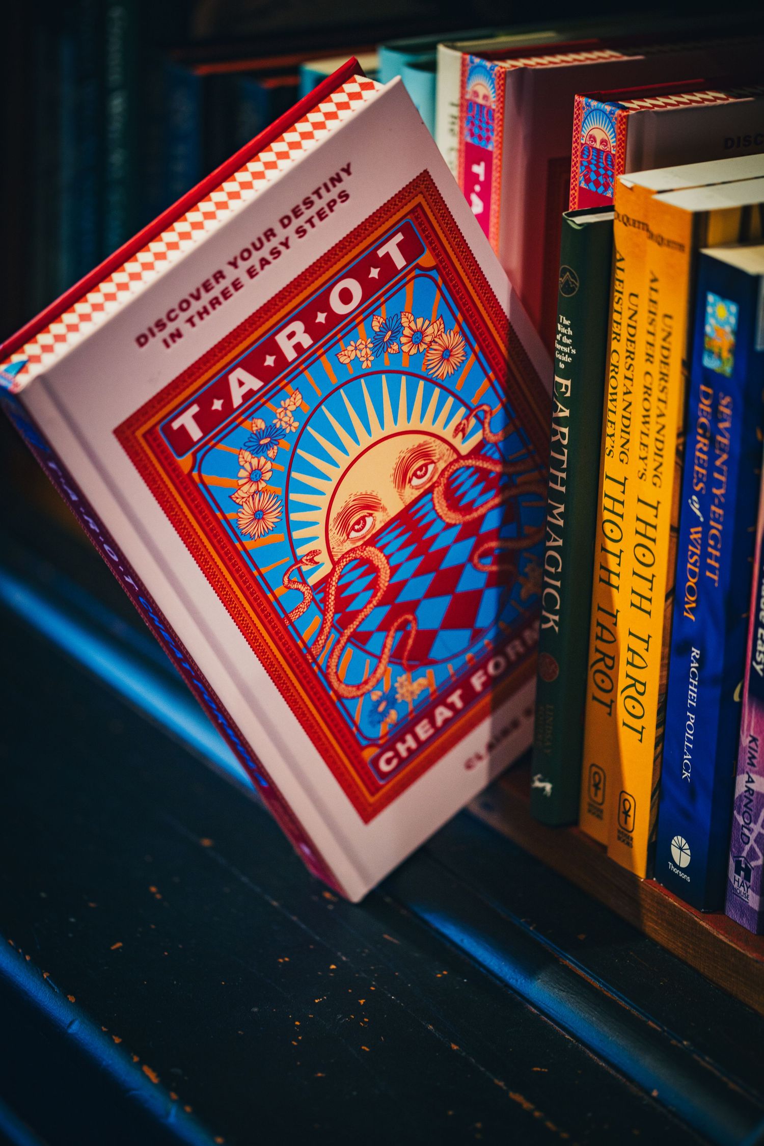 Colorful Tarot Cheat For Beginners book displayed among spiritual and mystical books on shelf.