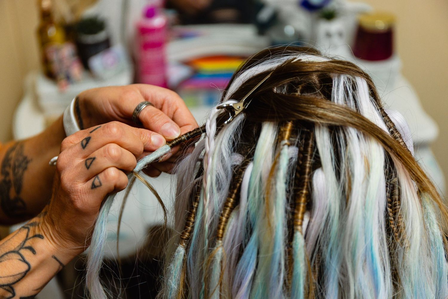 Close-up of tattooed hands installing colorful blue and gold dreadlock extensions into long hair.