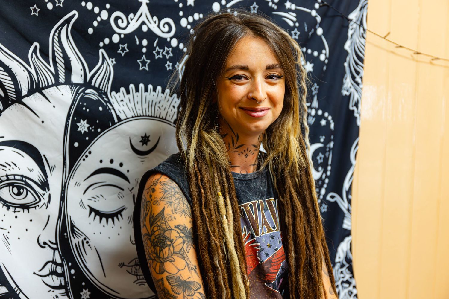 Woman with dreadlocks and tattoos smiling in front of a celestial sun and moon tapestry backdrop.