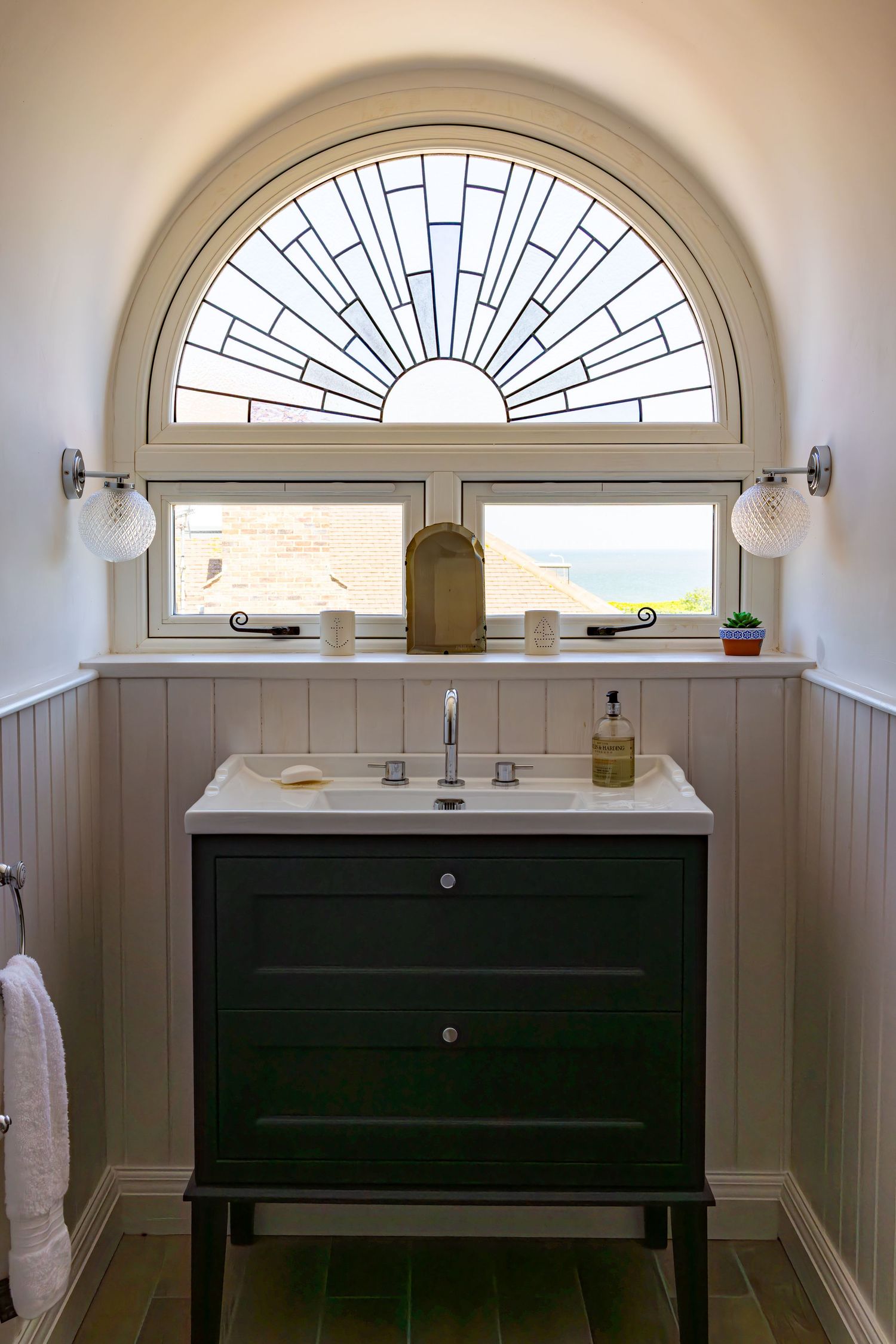 Elegant bathroom vanity with dark cabinet beneath arched Art Deco stained glass window with sea view.