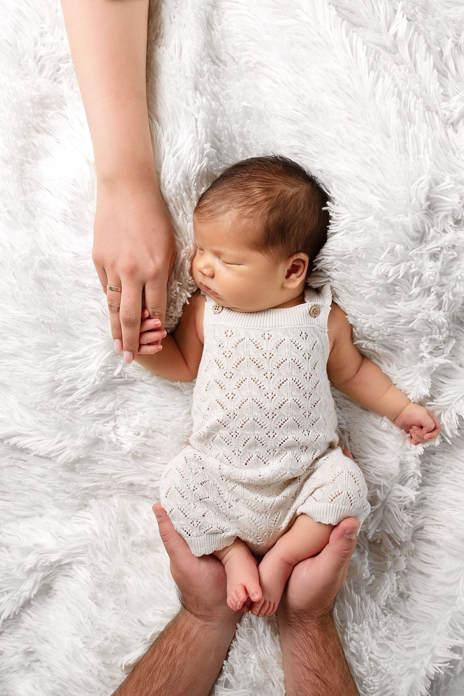 Newborn baby in cream knit romper sleeping on a white fluffy blanket held by parents.