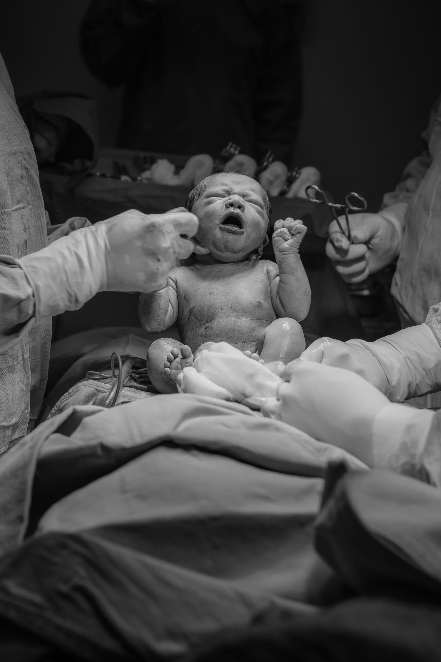 Newborn baby crying moments after birth, held by medical professionals in black and white delivery room photo.
