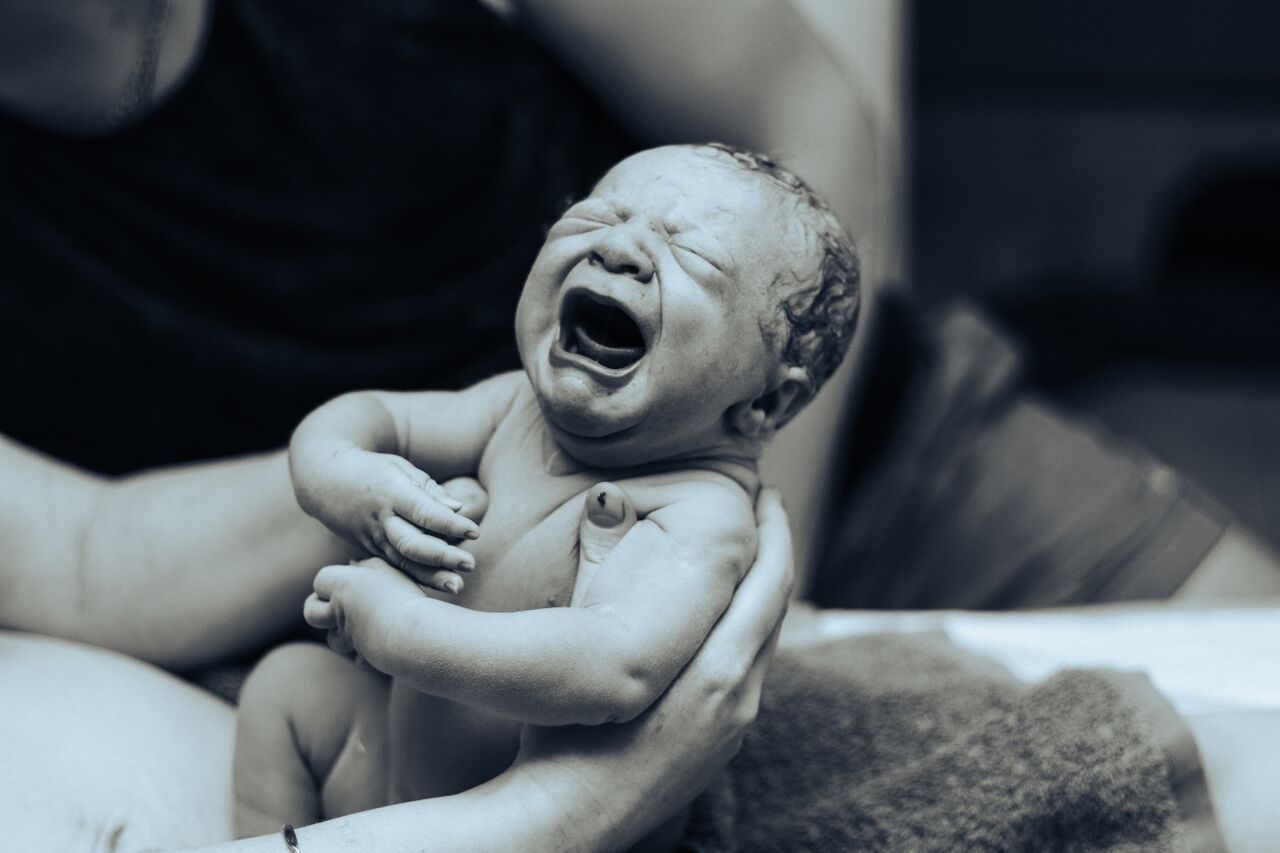 Black and white photo of a crying newborn baby being held by hands immediately after birth.