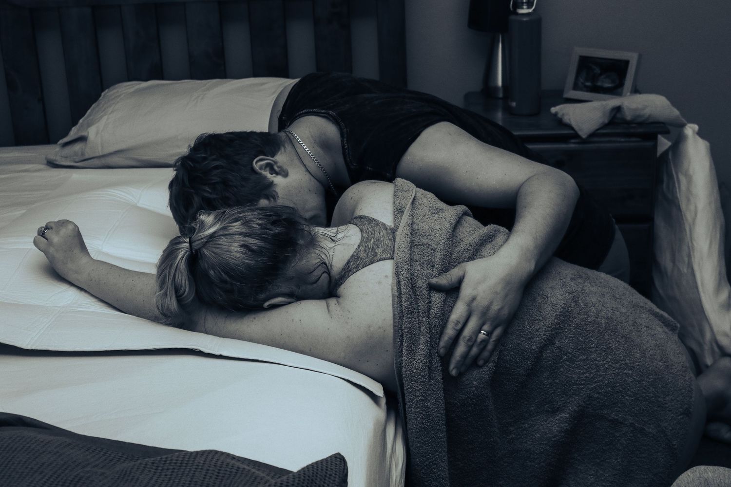 Couple lying together in bed in a tender embrace in a dimly lit bedroom.