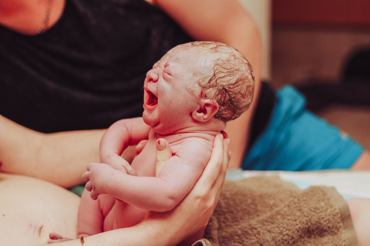 Newborn baby crying moments after birth, held in caring hands with umbilical cord still attached.
