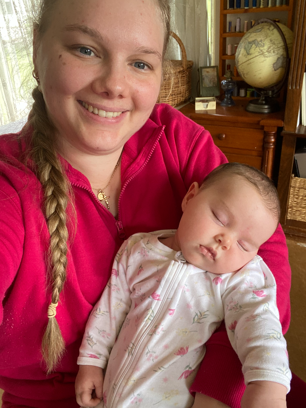 Smiling woman in pink hoodie with braid holds sleeping newborn baby in white floral onesie indoors.