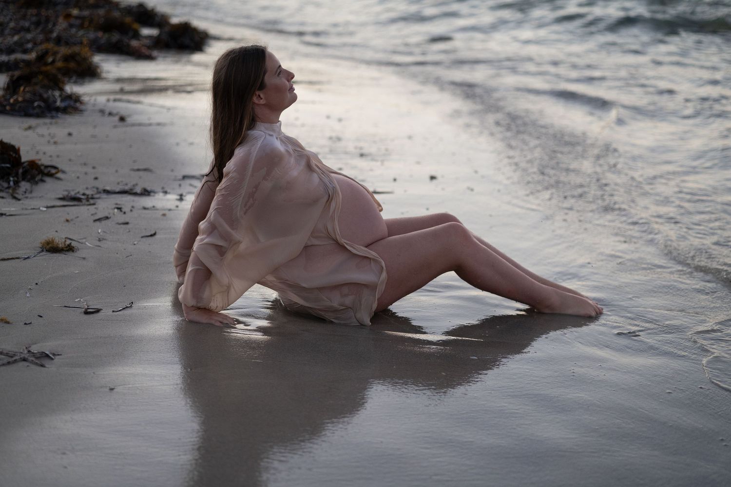 Pregnant woman in sheer white dress sitting peacefully on sandy beach shore with gentle ocean waves at dusk. Woodman Point Perth