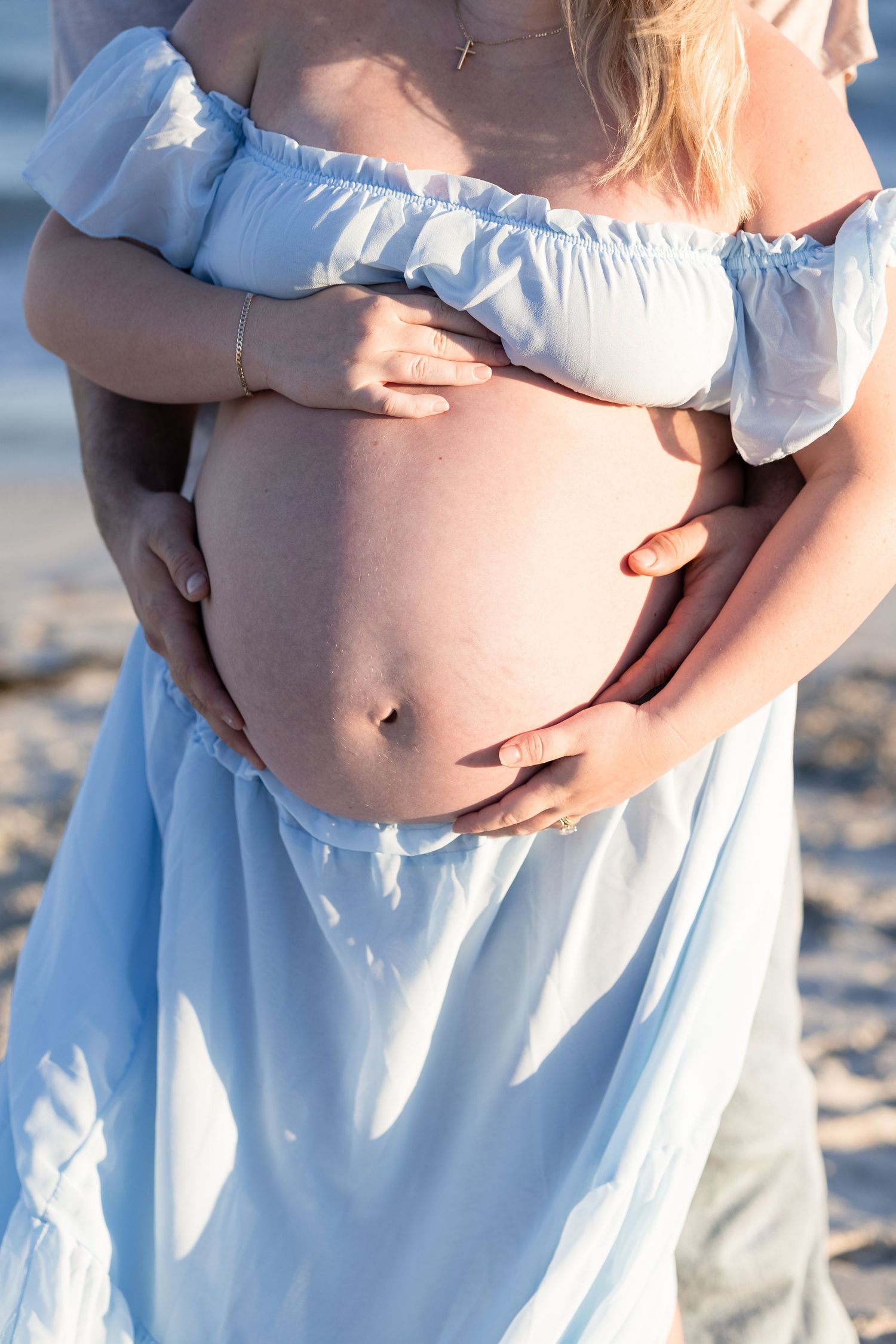 Pregnant woman in blue off-shoulder dress cradling baby bump on beach, partner's hands wrapped around her belly. Woodman Point Perth