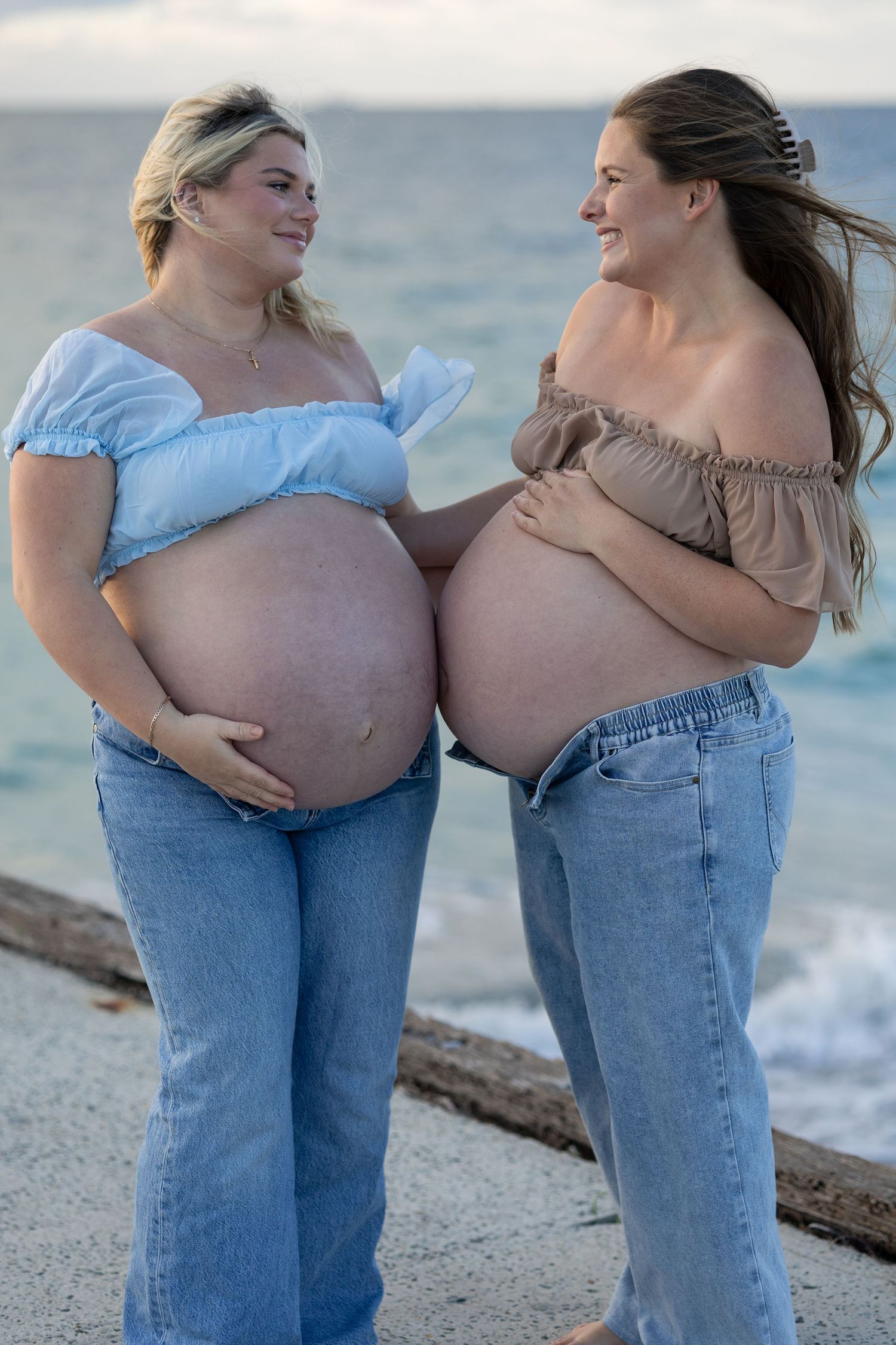 Two pregnant women in jeans and off-shoulder tops smiling at each other on a beach at sunset.