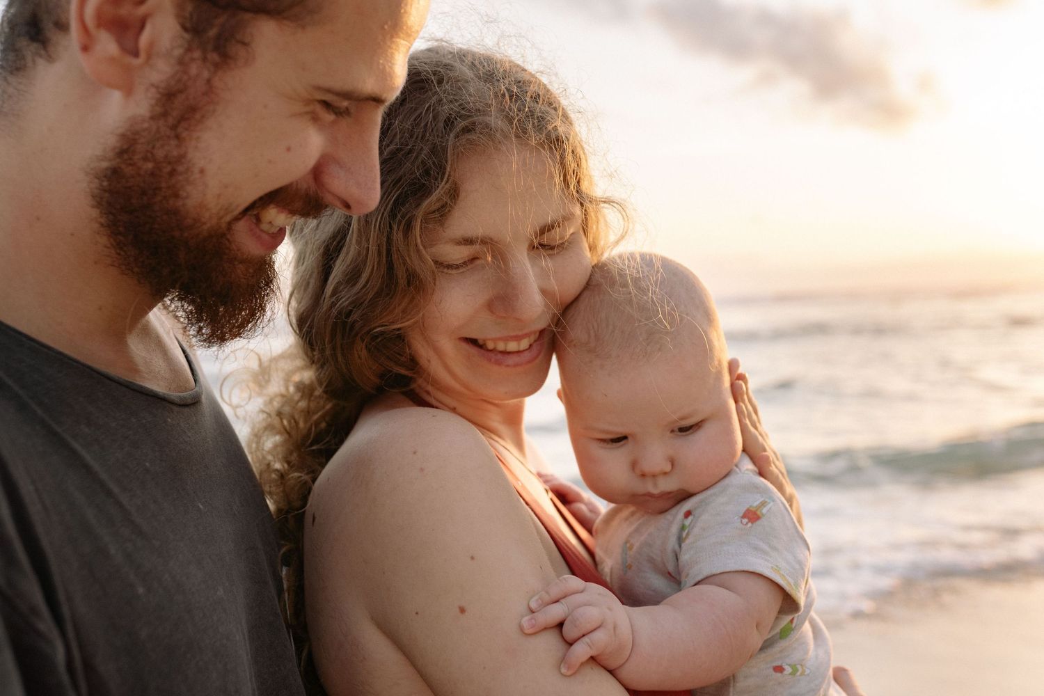Happy family of three with parents smiling at their baby on a beach at golden sunset.