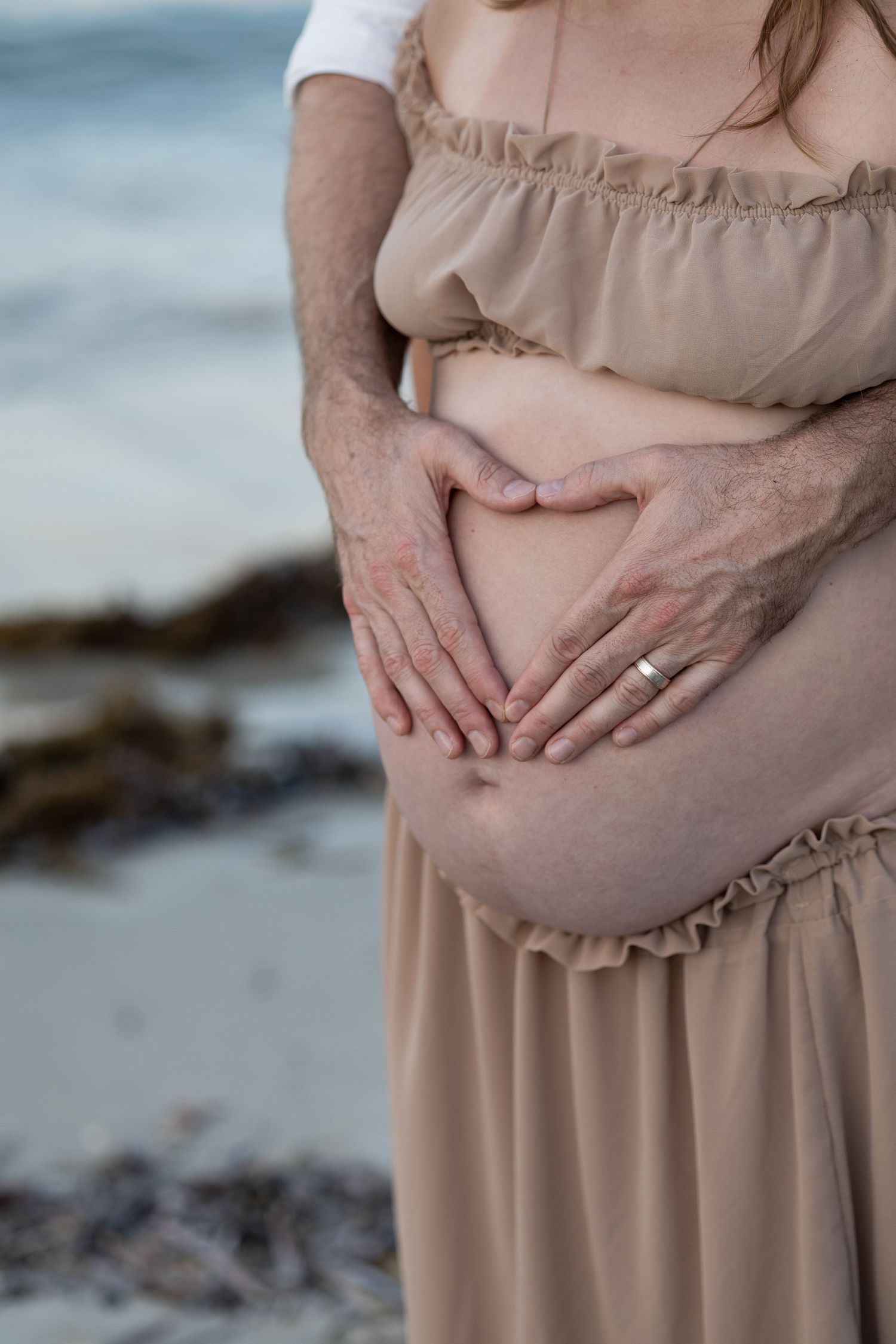 Couple forming heart shape with hands on pregnant belly at beach, woman in beige off-shoulder dress, ocean background.