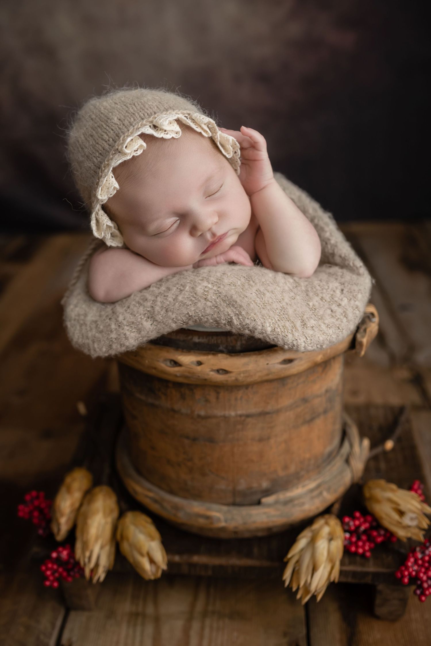 Sleeping newborn baby in a knit bonnet posed in a rustic wooden bucket with red berries.