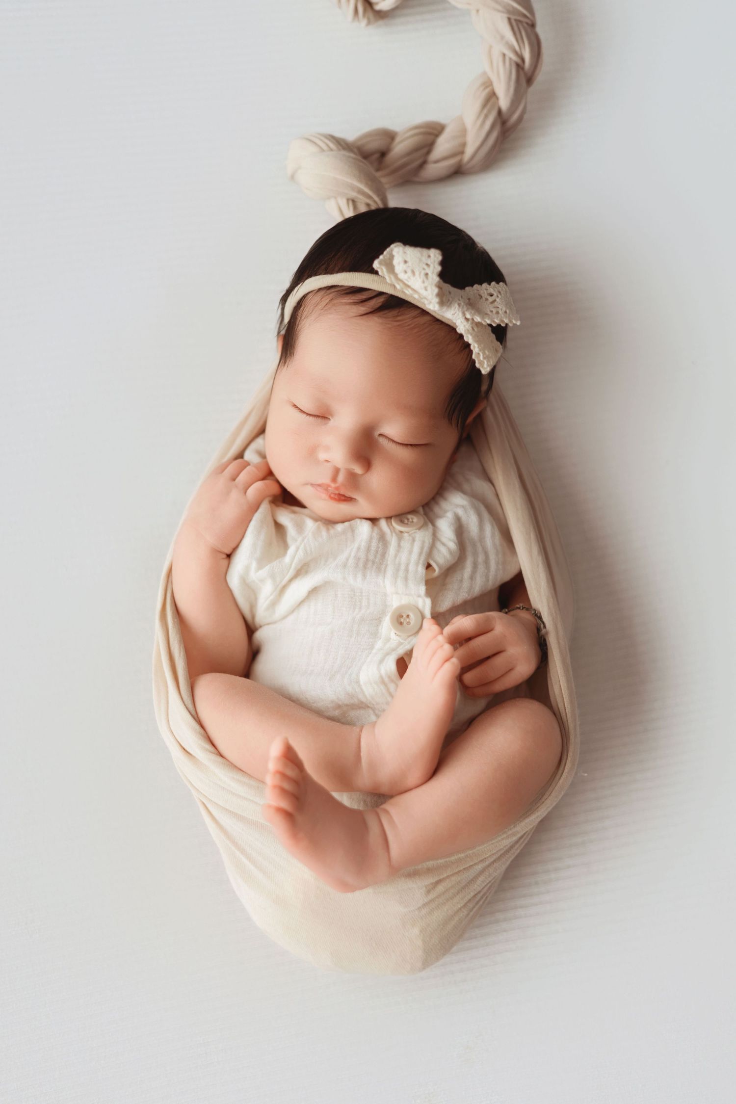 Sleeping newborn baby girl wrapped in cream fabric wearing a white bow headband on white background.