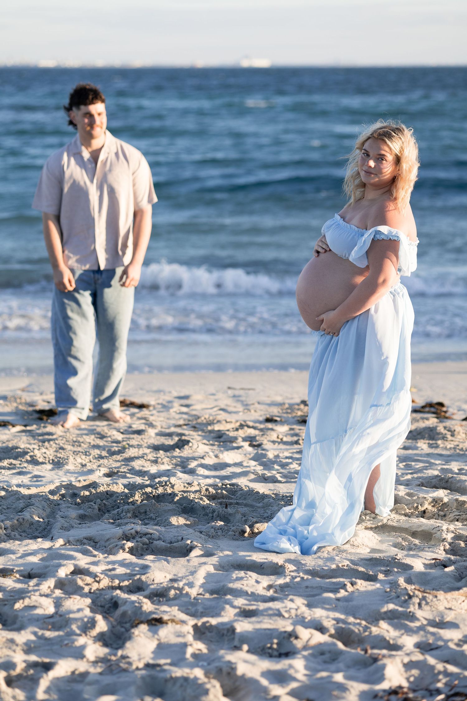 Pregnant woman in blue off-shoulder dress holds baby bump on beach while man stands behind her near ocean waves.