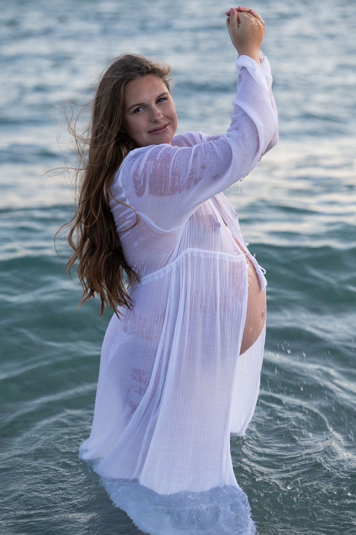 A woman in a wet white sheer dress stands in ocean waves, her long hair flowing in the breeze. Woodman Point Perth