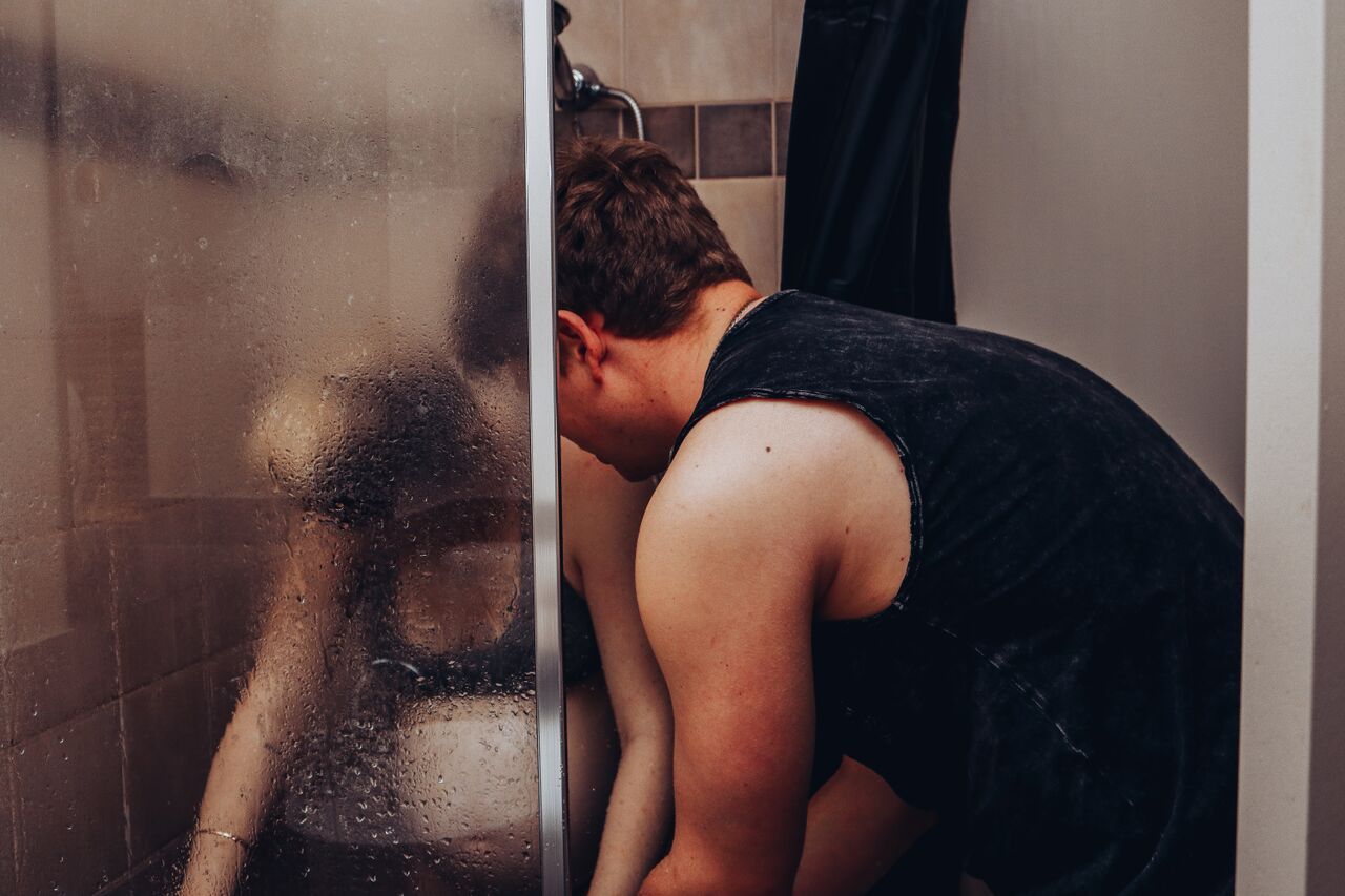 Person leaning into a steamy glass shower door in a dimly lit bathroom.