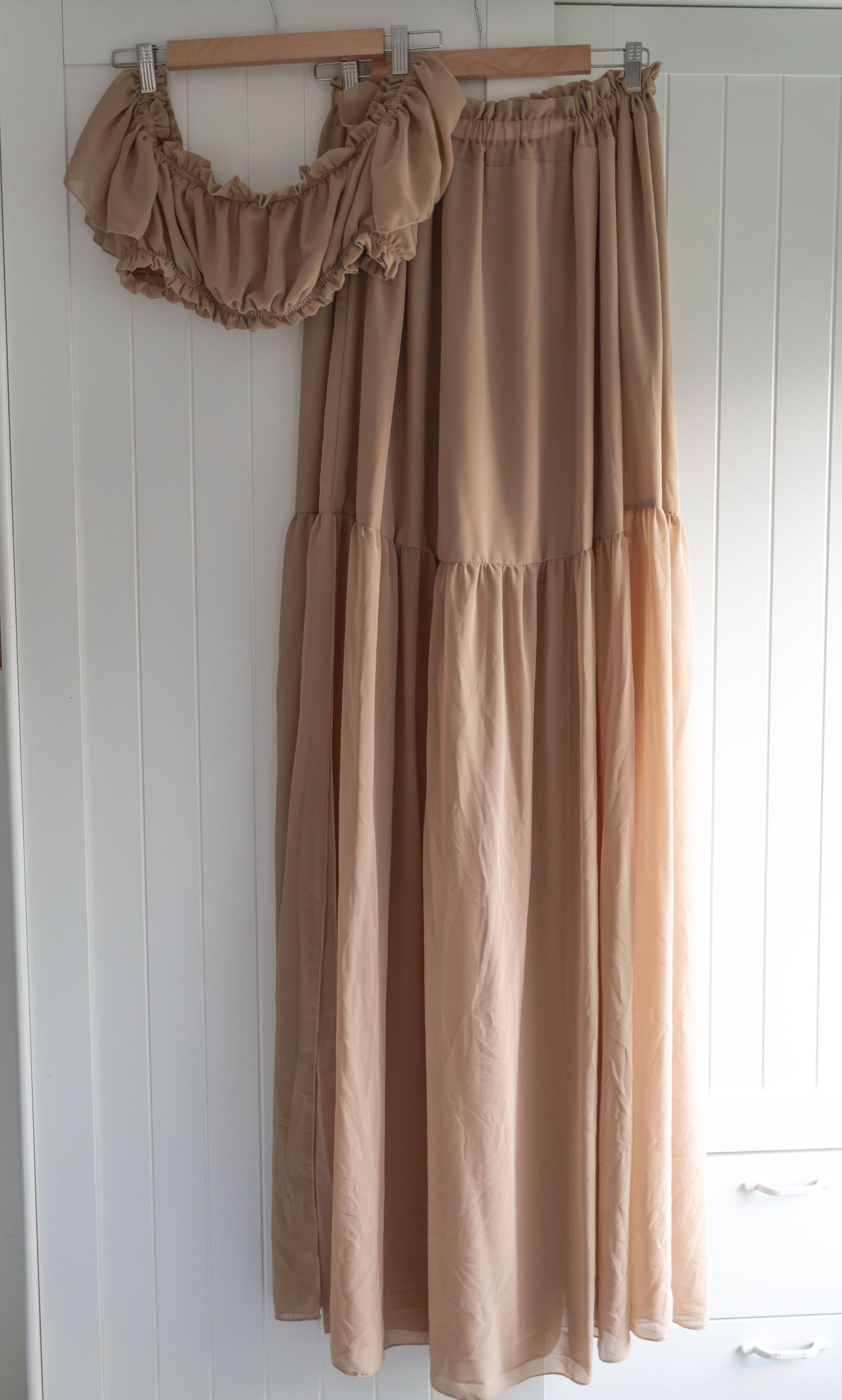 Beige off-shoulder ruffled top and matching maxi skirt two-piece set hanging on a wooden hanger.