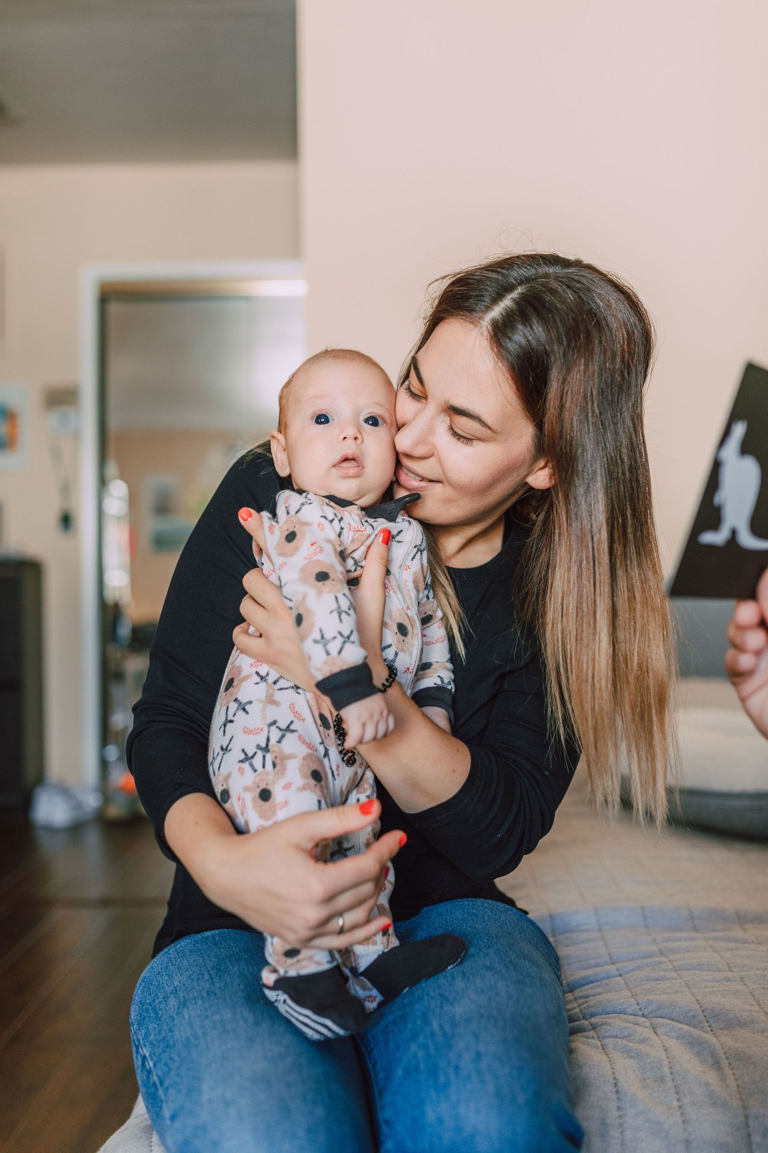 Young mother kissing smiling baby dressed in floral onesie while sitting on couch at home.