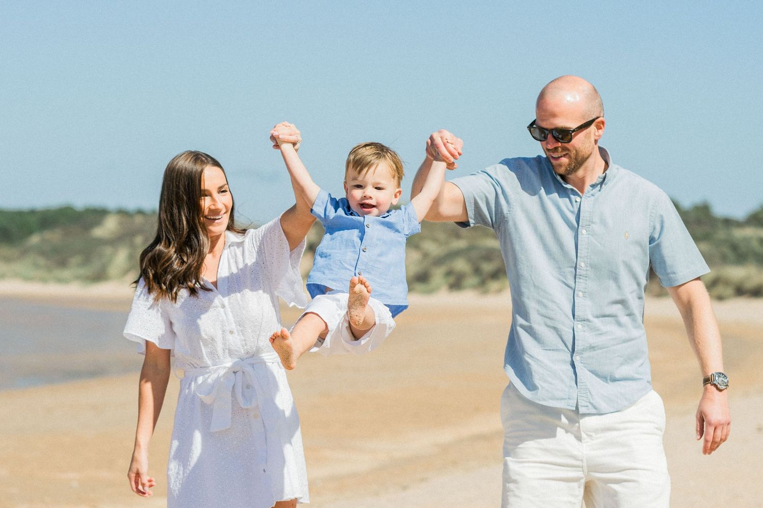 Joyful parents lifting their toddler son on a sunny beach with clear blue skies.
