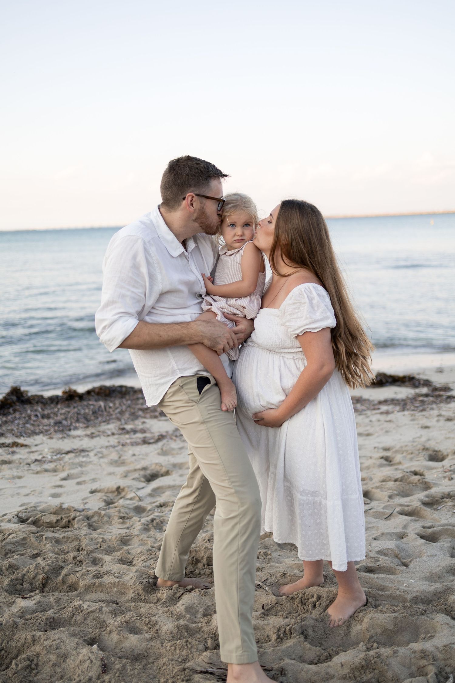 Pregnant couple kissing their toddler on a sandy beach at sunset, family maternity photo session. Taken by Golden Hour Perth at Cape Peron