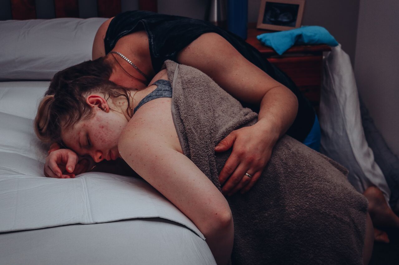 Two people sharing a tender embrace while resting on a bed in a softly lit bedroom.