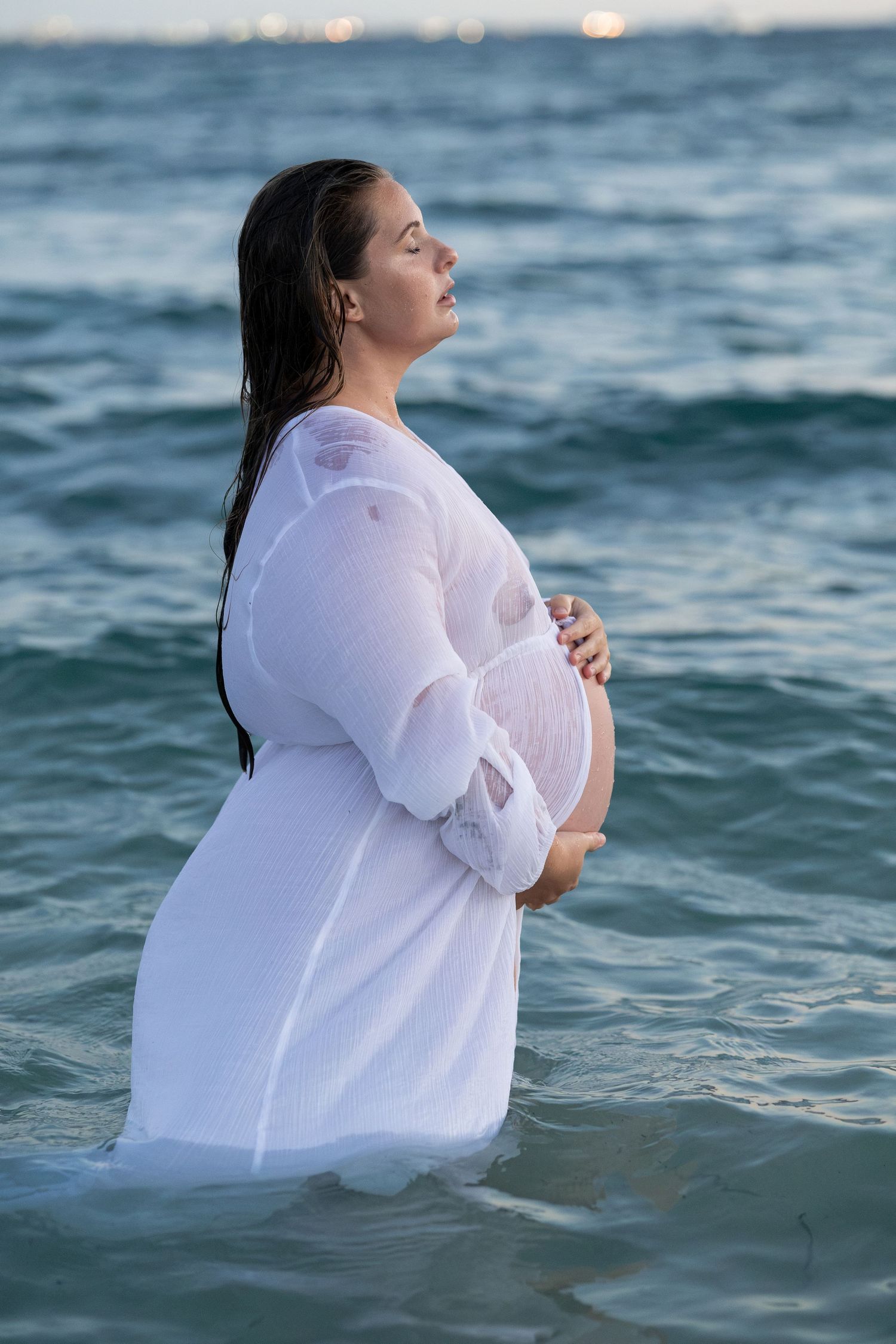 Pregnant woman in white dress standing in ocean waves, cradling her baby bump during a maternity photoshoot.
