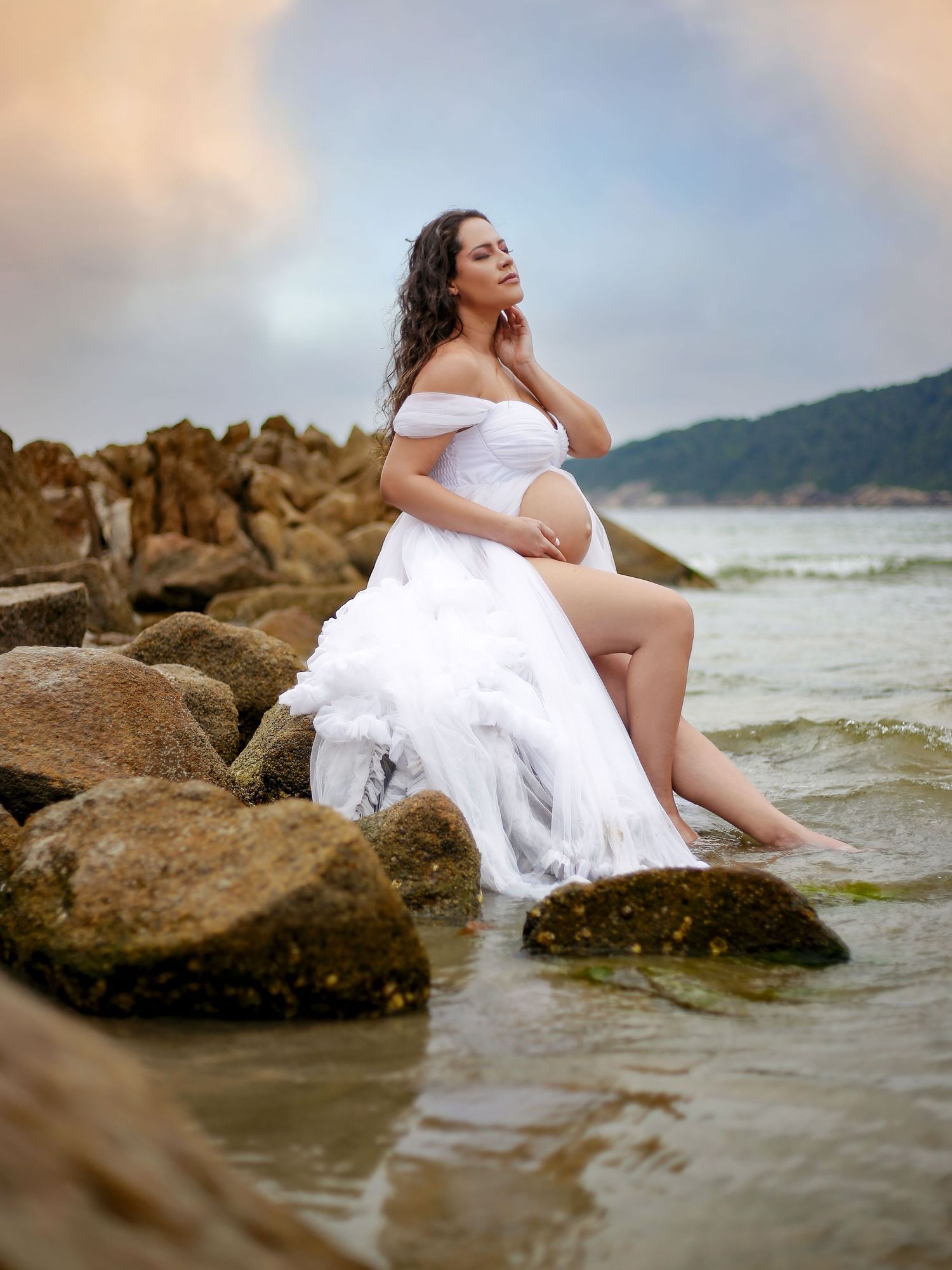 Pregnant woman in white off-shoulder gown sitting on coastal rocks with feet in water at sunset.