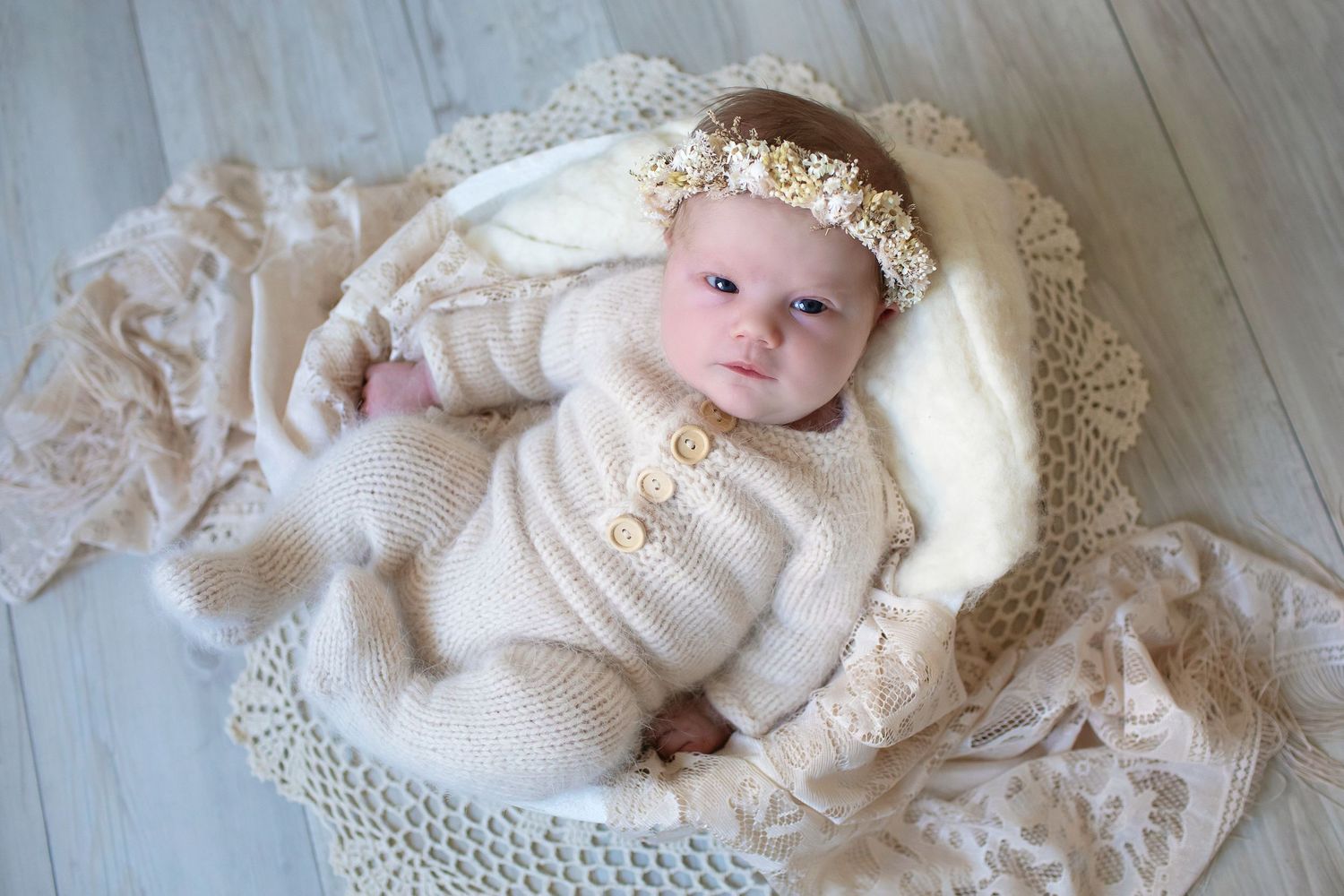 Awake newborn baby in beige knit outfit and floral headband lying on a crochet blanket.
