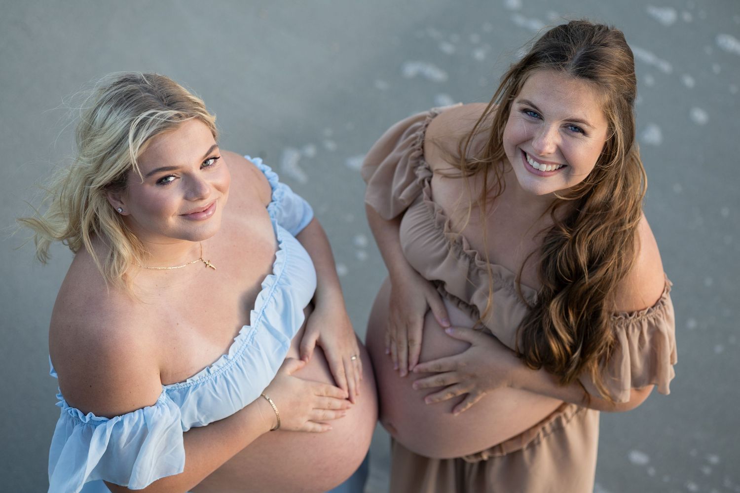 Two smiling pregnant women in pastel off-shoulder dresses pose together outdoors, cradling their baby bumps.