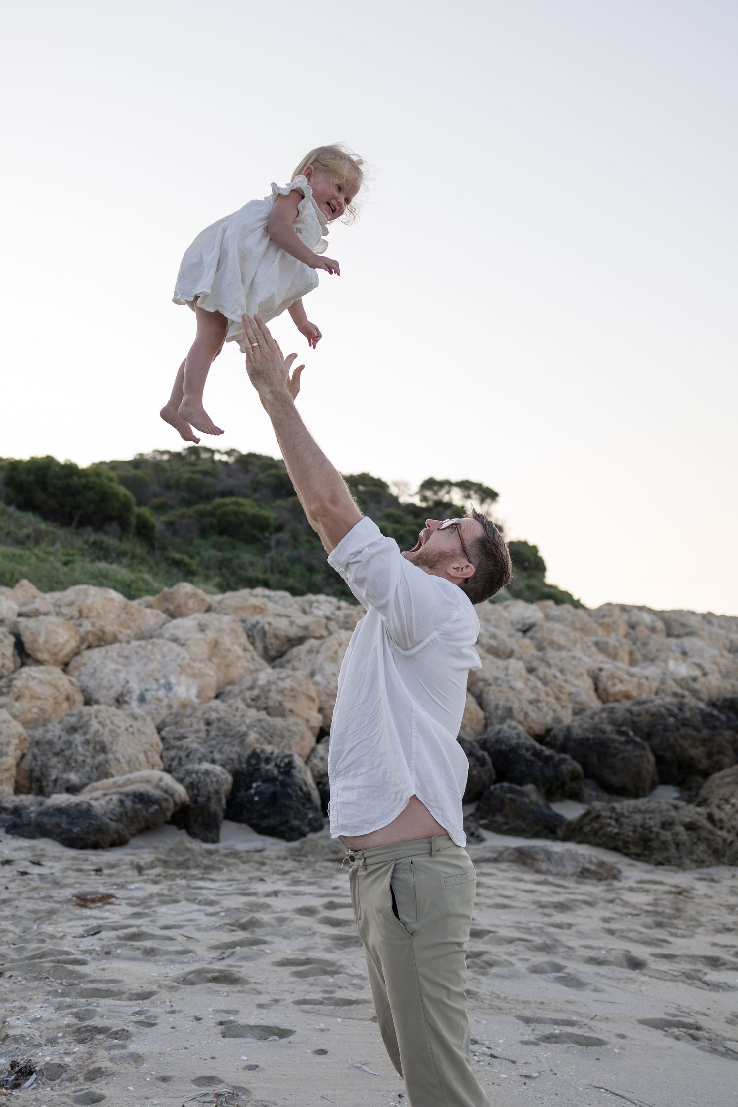 Father joyfully tossing young child in the air on a rocky beach, both dressed in white clothing. Cape Peron Perth