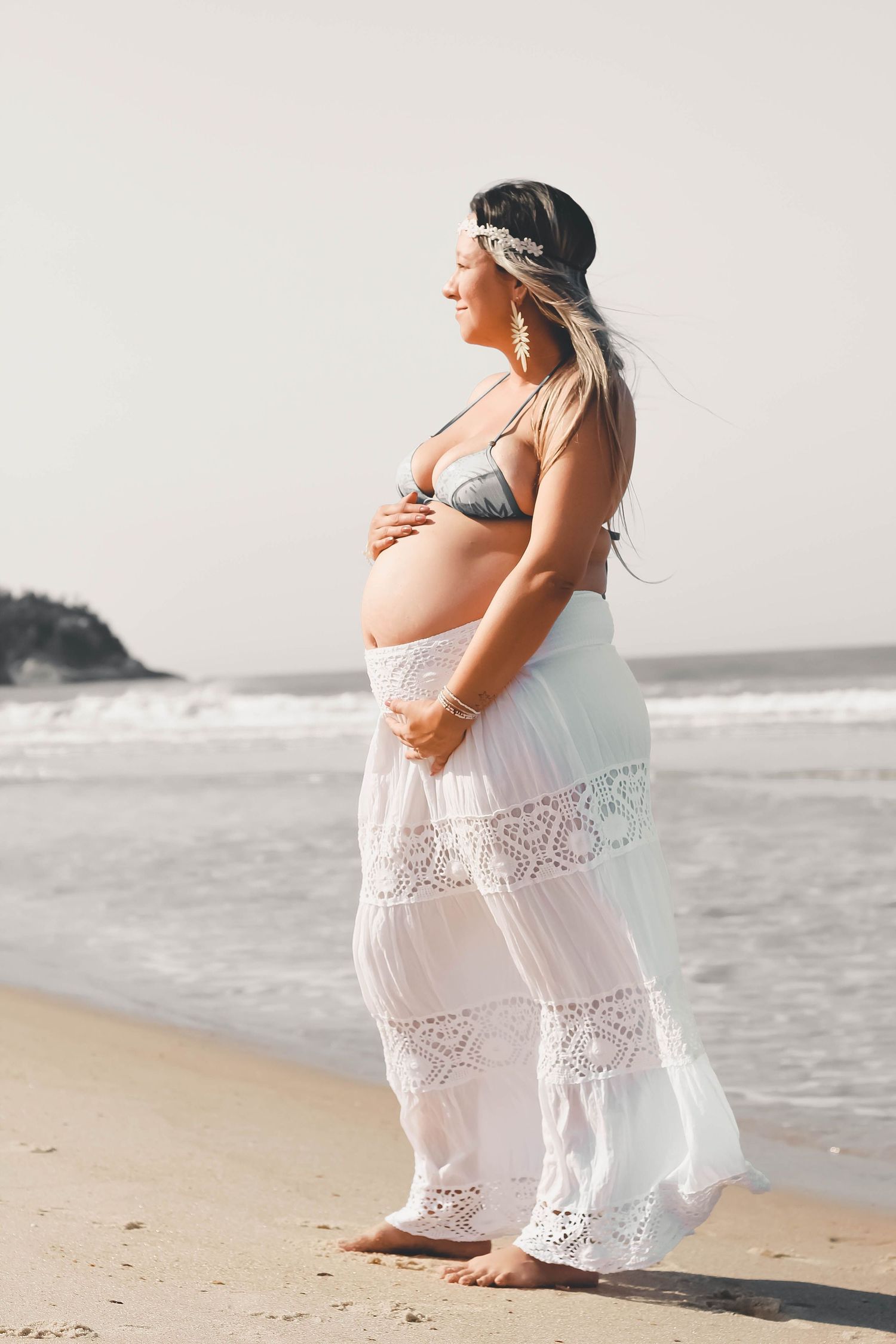 Pregnant woman in bohemian bikini and white lace skirt holding belly on sandy beach shoreline.