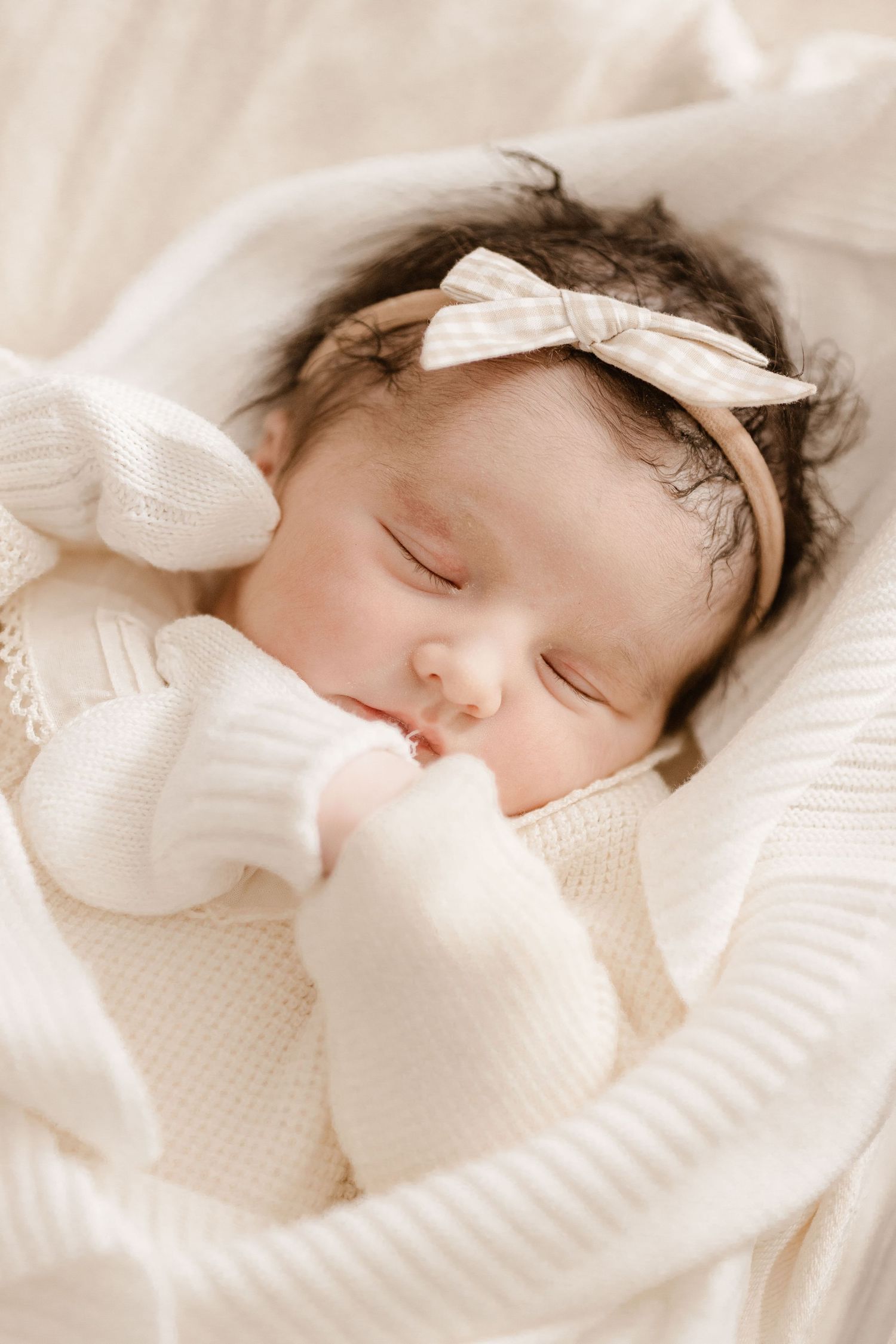 Sleeping newborn baby girl in white knit outfit with gingham bow headband nestled in soft blankets.