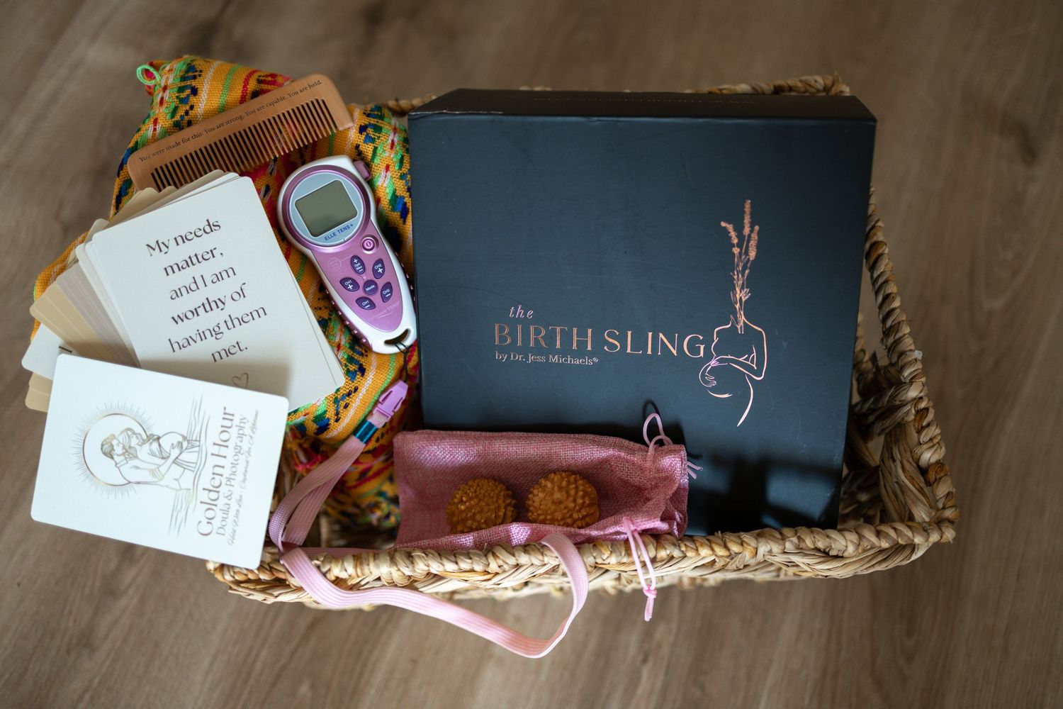 A wicker gift basket containing The Birth Sling box, a TENS machine, affirmation cards, and pink accessories for new mothers.