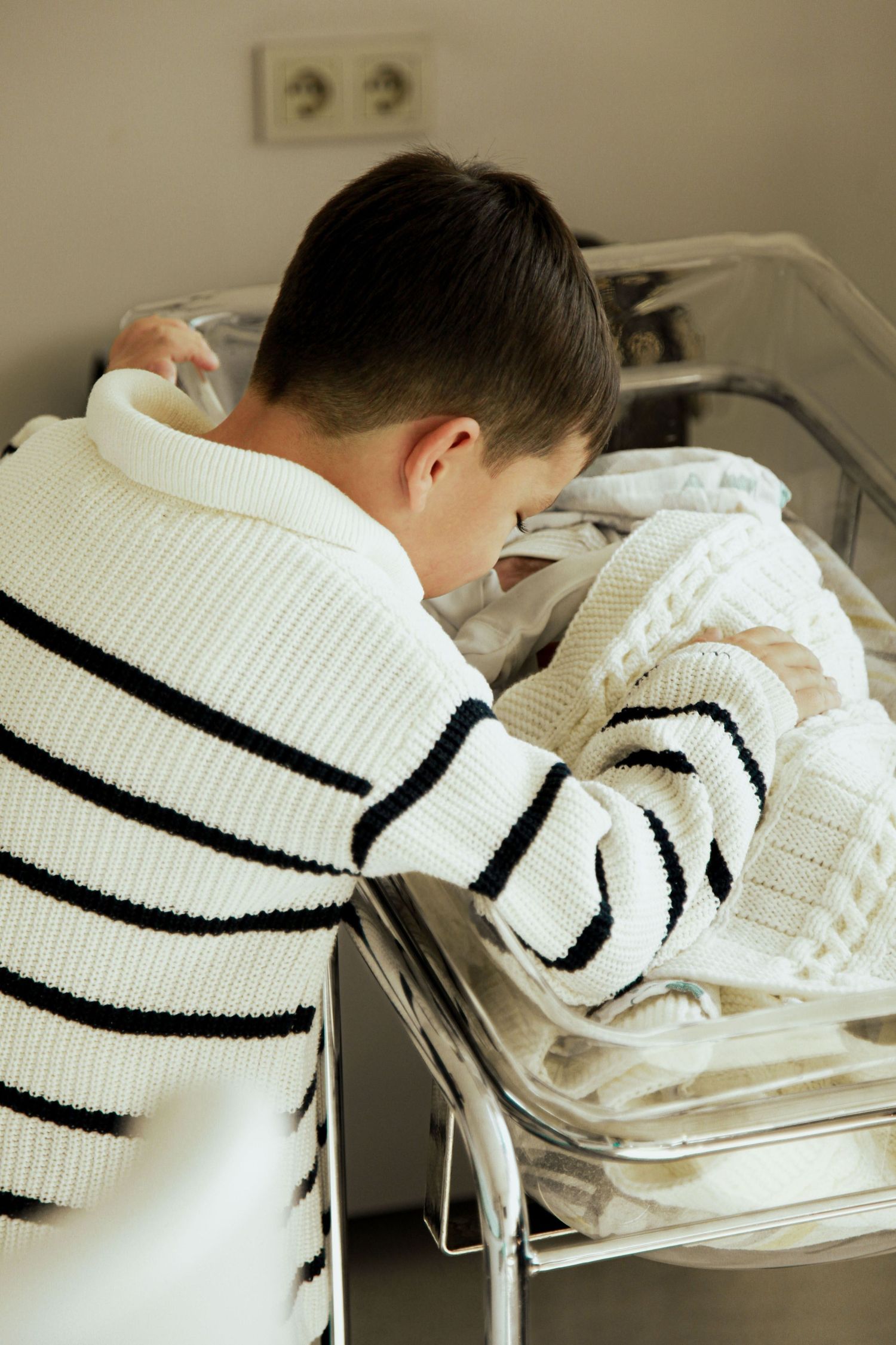 Young boy in striped sweater leaning over hospital bassinet to look at his newborn sibling.