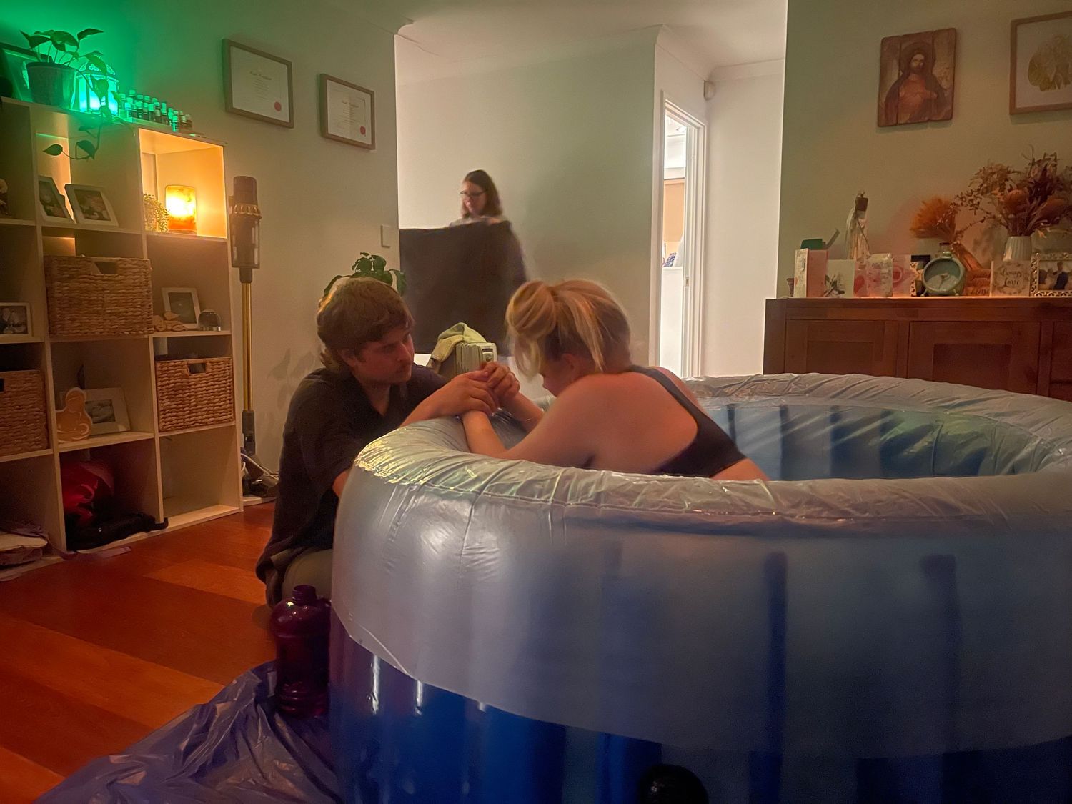 Two people leaning over an inflatable birthing pool during a home water birth in a cozy living room.