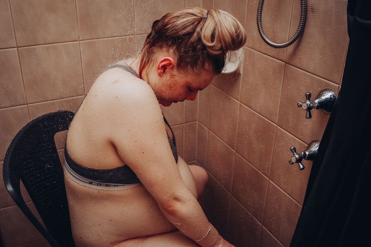 Woman sitting in a shower chair leaning forward in a tiled bathroom.