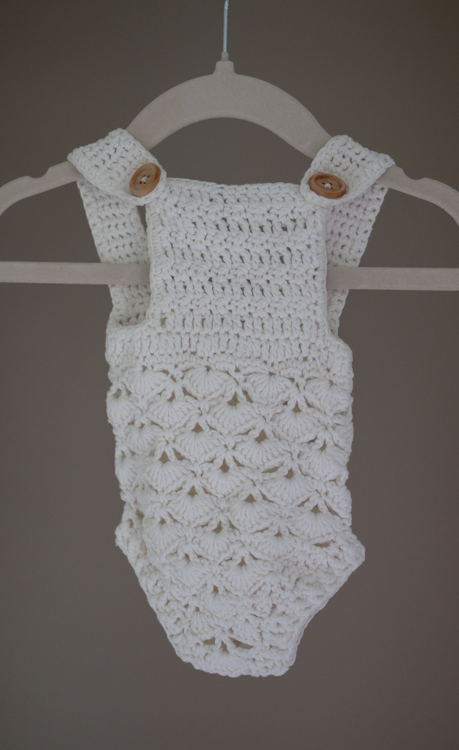 White crochet baby romper with shell stitch pattern and wooden buttons on straps, hanging on a hanger. Taken by Golden Hour Perth