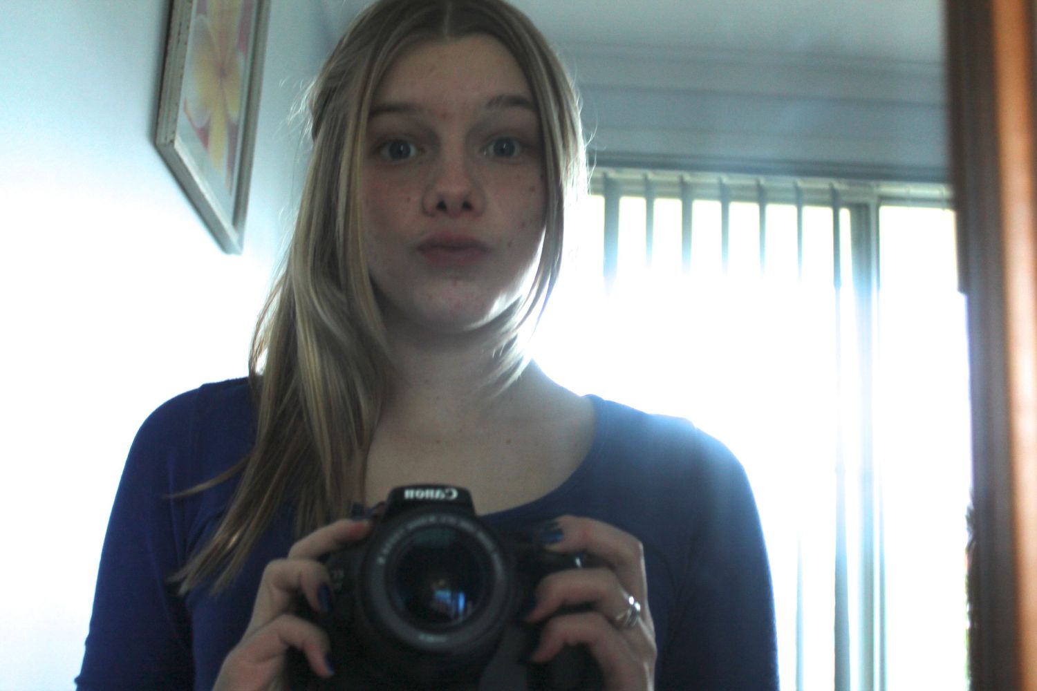 Young blonde teenage girl taking mirror selfie with Canon DSLR camera wearing blue top indoors.