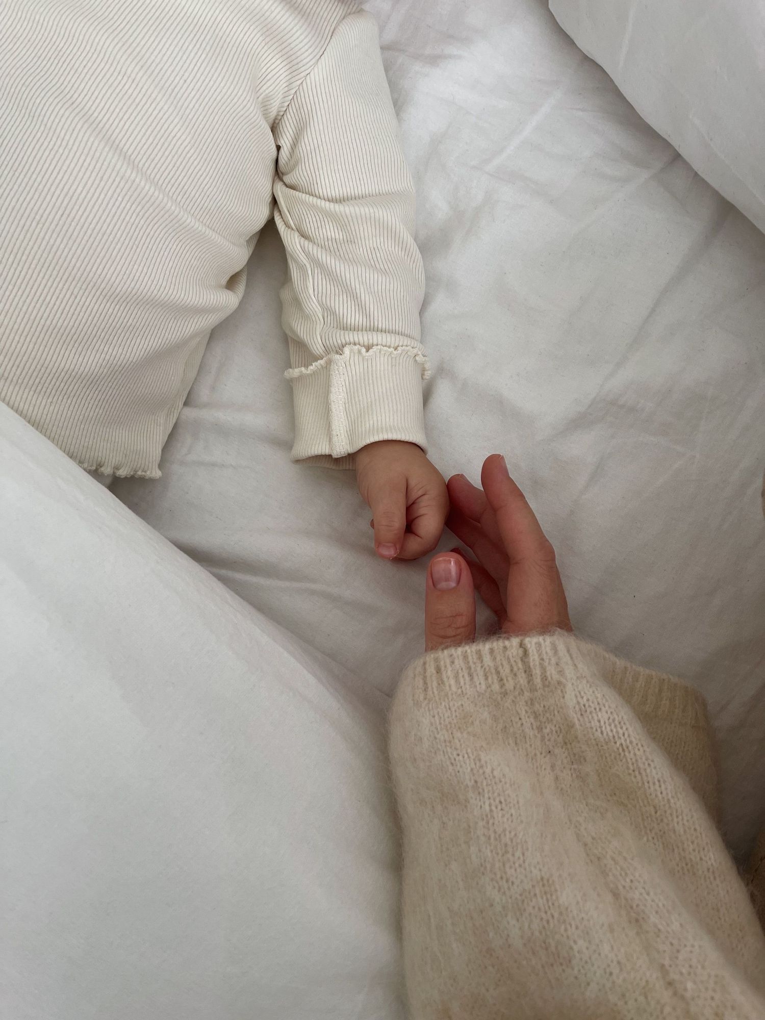 Parent holding tiny newborn baby hand wearing cream outfit on white bedding.