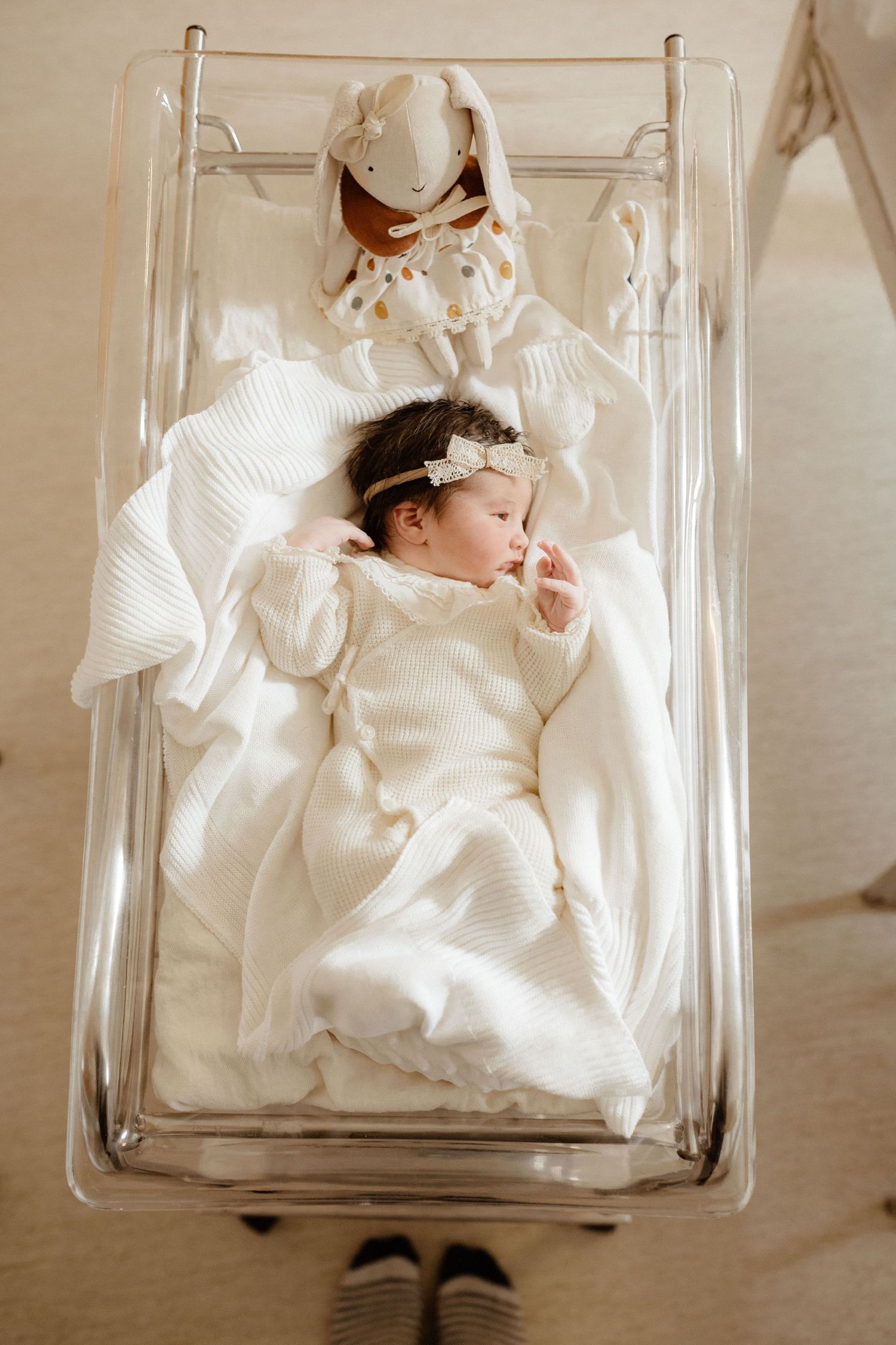 Overhead view of sleeping newborn baby in white outfit and headband in a clear hospital bassinet.