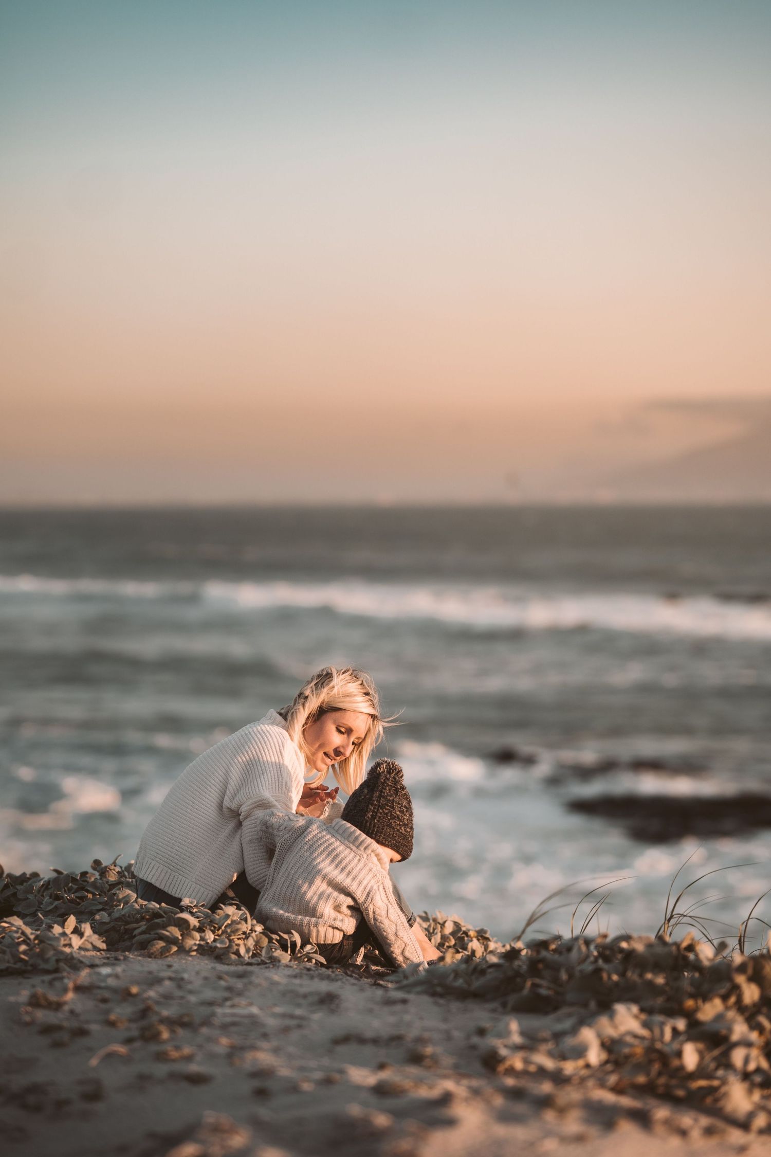 Blonde woman sitting on rocky beach at sunset in white sweater looking down peacefully.